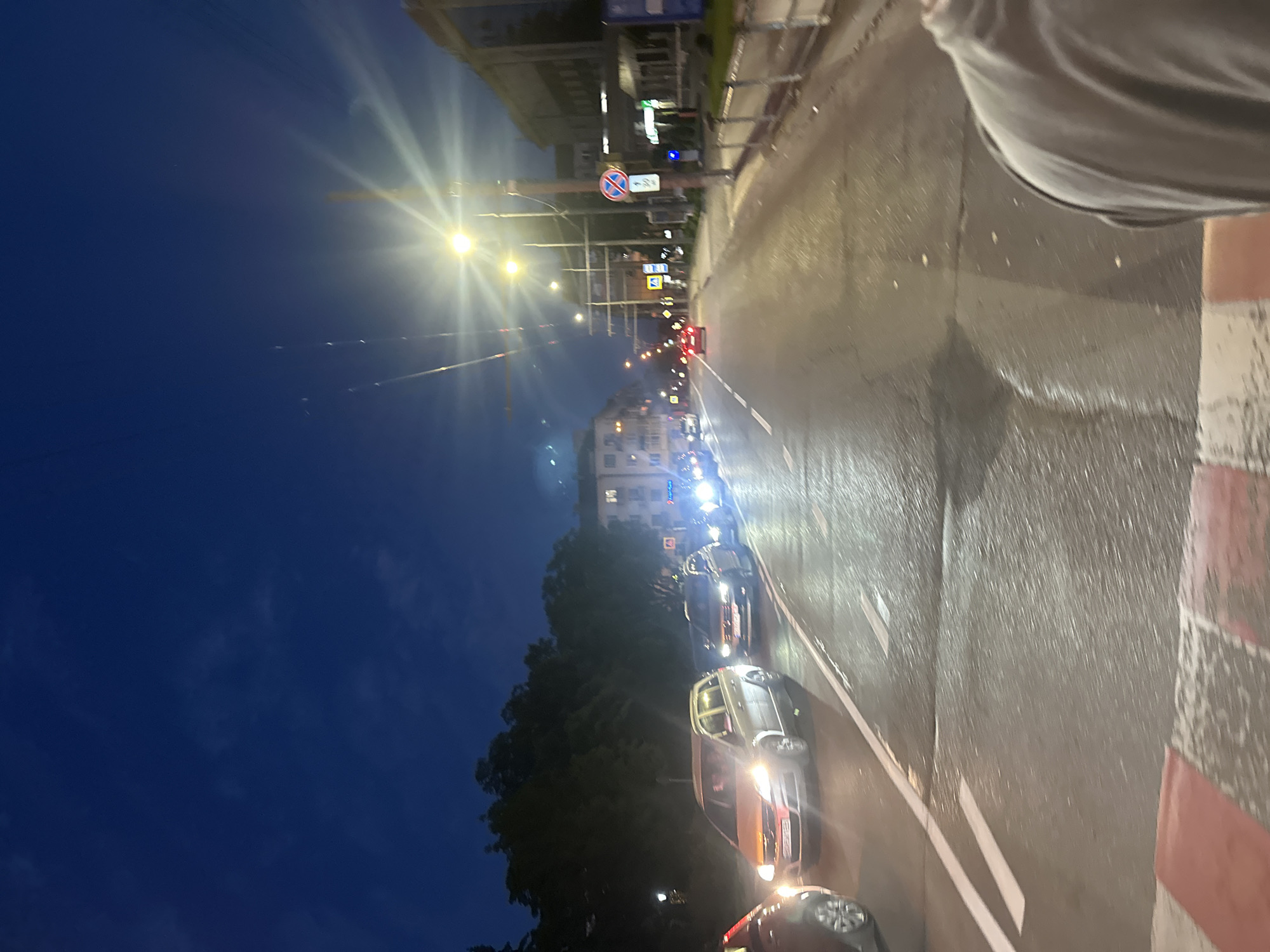 The image depicts a nighttime urban street scene in Ternopil, Ukraine. The sky is a deep, dark blue with faint cloud formations. The wet asphalt road surface reflects the bright glow of numerous vehicle headlights and streetlights, creating visible light streaks and reflections. In the foreground, the bottom right quadrant reveals the upper shoulder and back of a person, indicating the photograph was taken from a first-person perspective, likely by someone standing at the edge of the road or behind a low barrier. A red and white striped barrier, possibly a temporary construction barrier or painted curb marking, is partially visible at the very bottom. The street is a multi-lane thoroughfare with a double white line separating opposing directions of traffic. The left lane shows a line of cars, including a silver station wagon (likely an Opel Zafira or similar) prominently positioned, with its headlights illuminating the wet road ahead. The visible partial license plate on this silver car starts with "BO", which is the regional code for Ternopil Oblast in Ukraine, confirming the general location. Other vehicles, mostly sedans and hatchbacks, are visible further down the left lane and in the right lane, all with their headlights on, some showing red taillights. A red car is distinctly visible further down the road on the right lane. To the left of the road, a dense line of dark trees forms a natural barrier, obscuring any structures behind them. Overhead, multiple trolleybus wires crisscross above the street, indicative of public transportation infrastructure. On the right side of the street, a row of multi-story buildings with various architectural details lines the sidewalk. Some ground-floor sections appear to house businesses, although specific signage is unreadable due to distance and blur. Several bright streetlights with prominent starburst lens flares illuminate the street and sidewalk. Multiple utility poles are present, supporting the overhead wires and various street signs. One pole prominently displays a blue circular sign with a red 'X' and a diagonal red line (indicating "No Stopping"), with a smaller white rectangular sign mounted below it containing unreadable Ukrainian text. Further down the street, a yellow diamond-shaped sign (indicating a "Priority Road" in Ukrainian traffic regulations) is also visible. Metal barriers or fences run along the sidewalk, separating pedestrians from the road. The scene conveys a sense of moderate to heavy traffic flow during the evening or night, possibly after rain, on a main