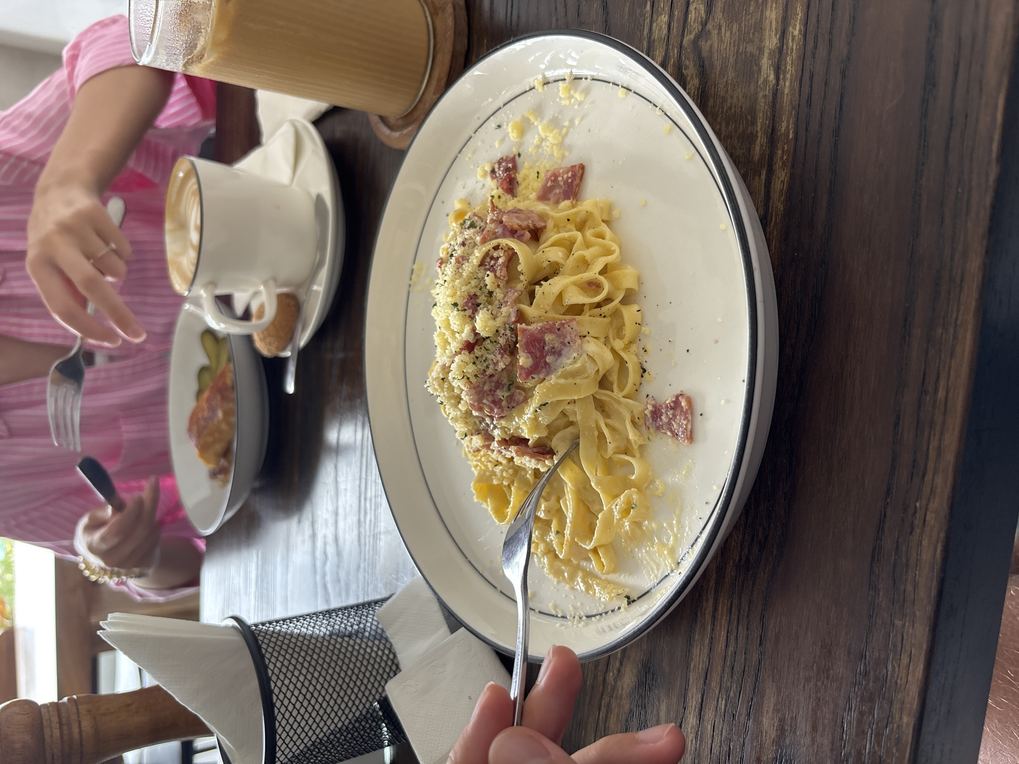 The image depicts a person eating carbonara pasta with a fork from a white plate with a black rim. The pasta is topped with cheese and bits of bacon. Adjacent to the pasta is a cup of coffee with a saucer and a tall glass of a brown liquid, possibly juice or another type of coffee. Another person is seen in the frame, wearing a pink striped shirt and holding a fork. They are eating a separate meal on a white plate. There is also a napkin holder on the table. The setting is likely a restaurant or cafe. The image was taken indoors and the table is made of wood. It is not possible to confirm the city location without additional context.