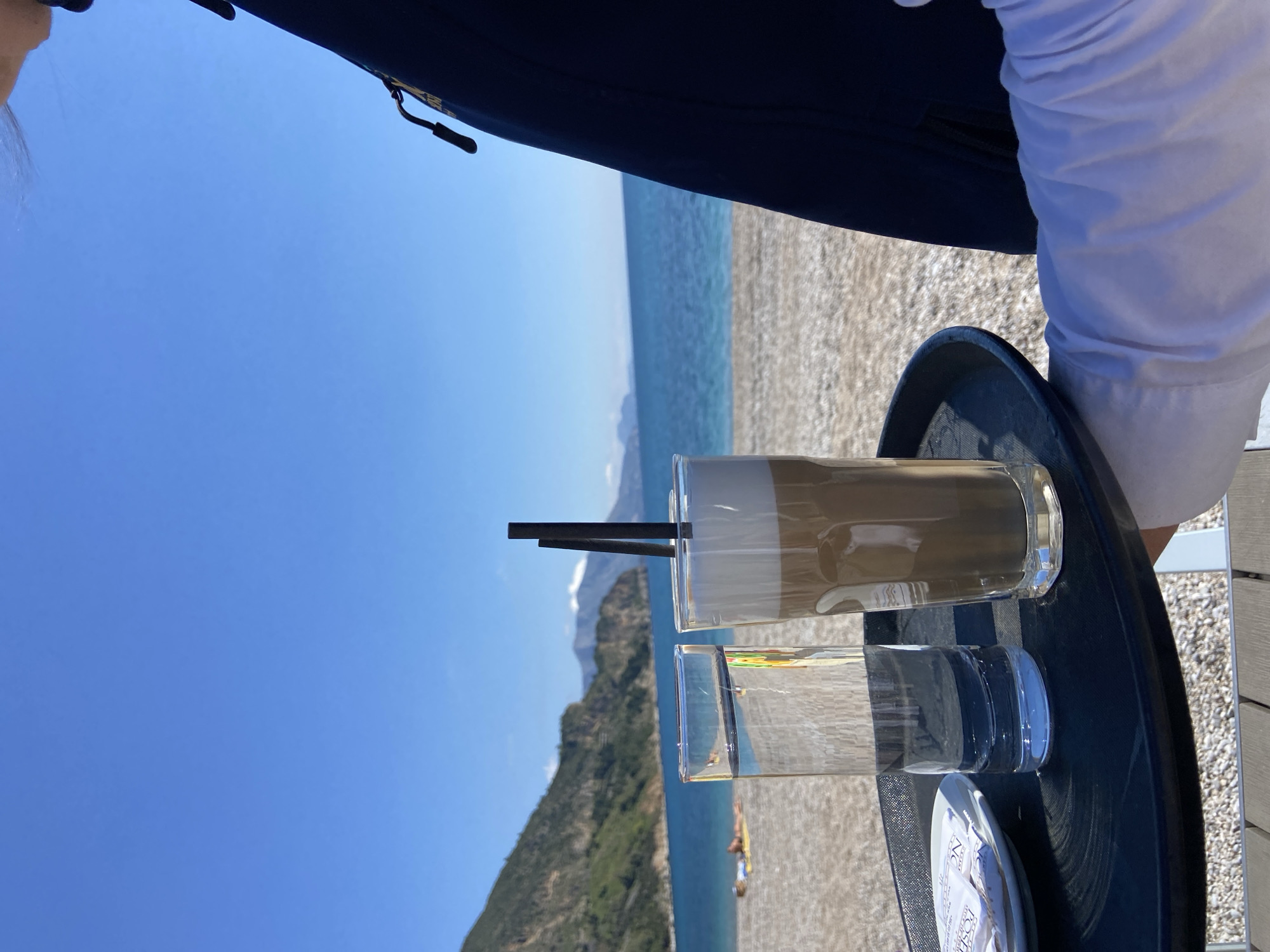 On a sunny day, a coffee drink and a glass of water sit on a black tray. The backdrop consists of a blue sky, a rocky beach, and the sea. Green hills are visible along the coastline. The scene is potentially located in or near Prijevor, Montenegro. A person wearing a white shirt is partially visible to the right. A dark-colored garment or object appears in the top-center to the left.