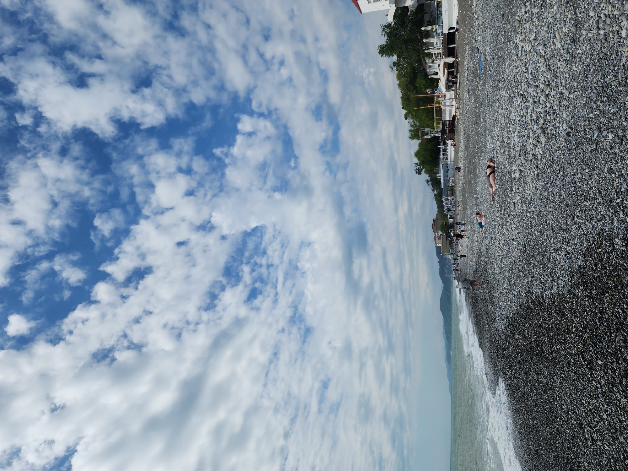 The image depicts a beach in Sochi, Russia. The beach is covered in pebbles and stretches along the coastline, where waves are breaking. Several people are present, including a woman lying down sunbathing and a person standing in the water. In the background, there are buildings, trees, and other beach structures like a volleyball net. The sky is filled with white clouds, providing a contrast to the blue sky.