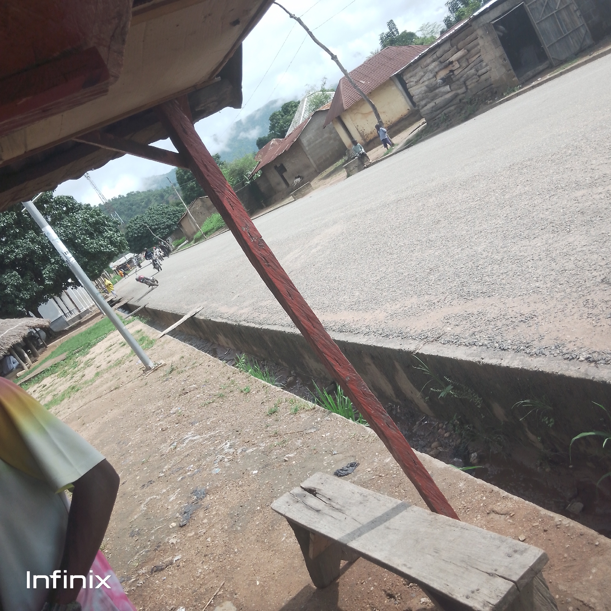An outdoor, eye-level, slightly tilted medium shot captures a roadside scene in an unspecified urban location. The foreground is dominated by the edge of a roofed structure with wooden support beams and a partial view of a person wearing a colorful shirt and skirt. A simple wooden bench sits on a dirt and gravel surface in the lower right quadrant. A shallow concrete drainage ditch runs alongside the paved road that occupies the middle ground. The road is sparsely populated with a few motorcycles and a person walking in the distance. Several buildings with corrugated metal roofs and walls constructed of various materials, including rough-hewn logs and concrete blocks, line the road on the right. In the background, lush green trees and misty mountains rise under an overcast sky. A tall pole is visible on the left side of the image. The overall impression is one of a rural or semi-urban settlement in a hilly, vegetated area.