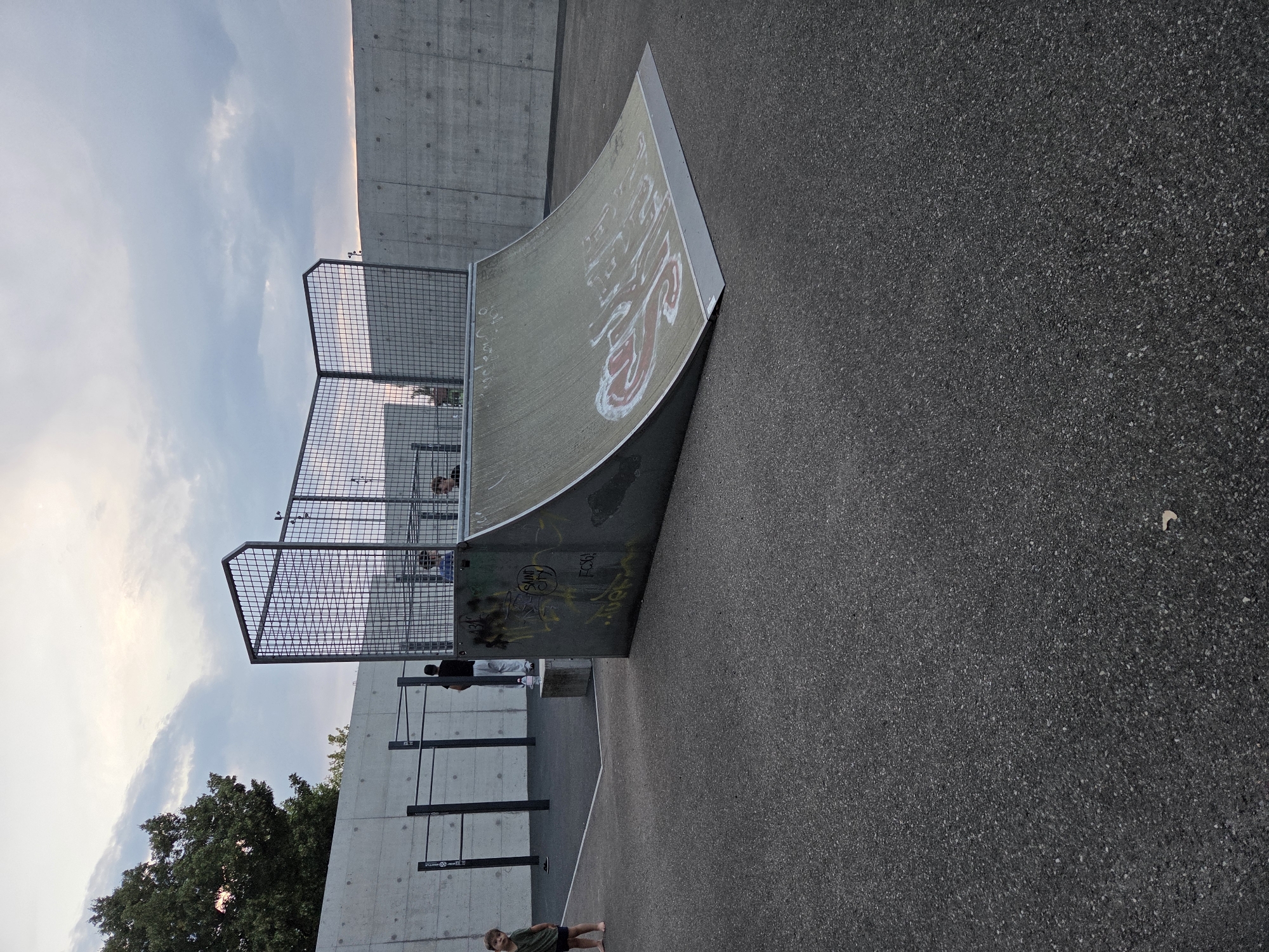 The image depicts an outdoor skate park facility, likely in Gossau (SG), Switzerland, given the context. In the foreground, a quarter pipe ramp with graffiti is visible. Behind it, a series of metal bars, possibly for calisthenics, are set up against a concrete wall. A wire mesh enclosure is situated behind the quarter pipe. Several individuals are present: a young child stands barefoot on the asphalt in the left foreground, facing the camera. A person in a dark t-shirt and white pants stands near the calisthenics bars. Two other individuals, appearing to be teenagers or young adults, are visible within the mesh enclosure, near the top of the quarter pipe. The environment includes a concrete wall and an asphalt ground surface. The sky above is partly cloudy.