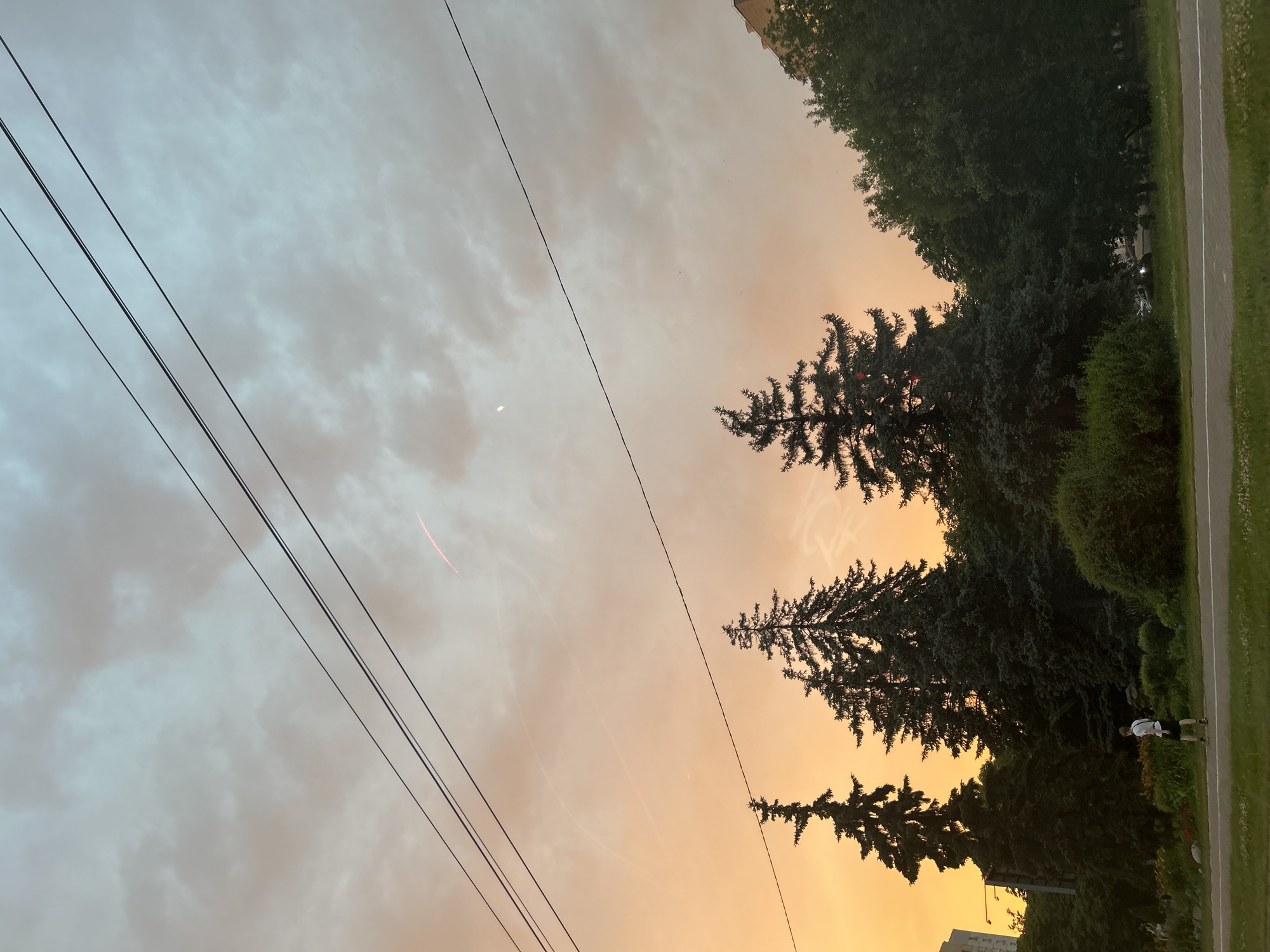 The image depicts a suburban street scene at dusk. The sky is a gradient of warm orange and soft blue hues, with scattered clouds and thin, faint contrails. Three parallel power lines stretch across the upper portion of the frame. In the foreground, a man wearing a white t-shirt and shorts is walking on the sidewalk, facing away from the camera. He is positioned in front of a dense line of evergreen trees. A paved road with a white line marking the edge runs in front of the sidewalk. Behind the trees, a residential building is partially visible on the right side of the image, and another taller building can be seen on the far left. The overall impression is of a quiet evening in a green, urban environment.