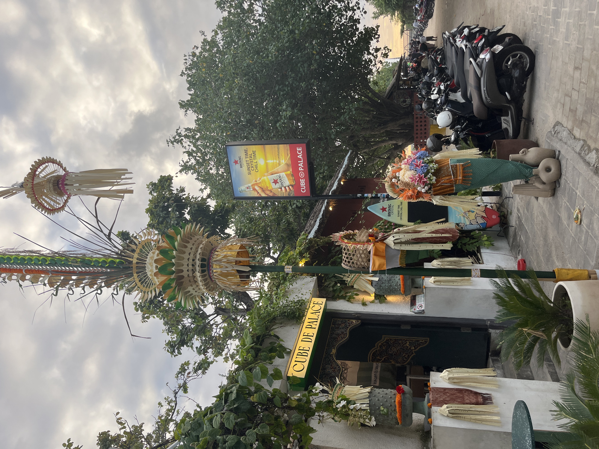 The image depicts an outdoor scene in Denpasar, Indonesia, likely near a beach or tourist area. In the foreground, a building with a sign that reads "CUBE DE PALACE" is visible, with a large green door and ornate carvings. Several decorative elements, including woven structures adorned with palm leaves and colorful fabric streamers, are placed outside the building. To the right of the building, a tall pole holds a large, intricate woven ornament, and another similar ornament hangs from a higher point.
A prominent billboard advertising "Bintang Crystal" beer with the slogan "Sunset time, Crystal chill time" is positioned next to a large tree with lush green foliage. The billboard features an image of a hand holding a bottle of beer against a sunset backdrop.
In the middle ground, a row of parked scooters and motorcycles stretches along a paved walkway. Several people are visible in the background, some walking and others standing near the parked vehicles. The ground is paved with cobblestones and some areas are covered with sand.
The sky above is partly cloudy, with the soft light suggesting it is either early morning or late afternoon. The overall impression is of a lively, possibly tourist-oriented establishment in a tropical setting. The presence of scooters and the beach-like advertisement suggest a location where tourists commonly visit.