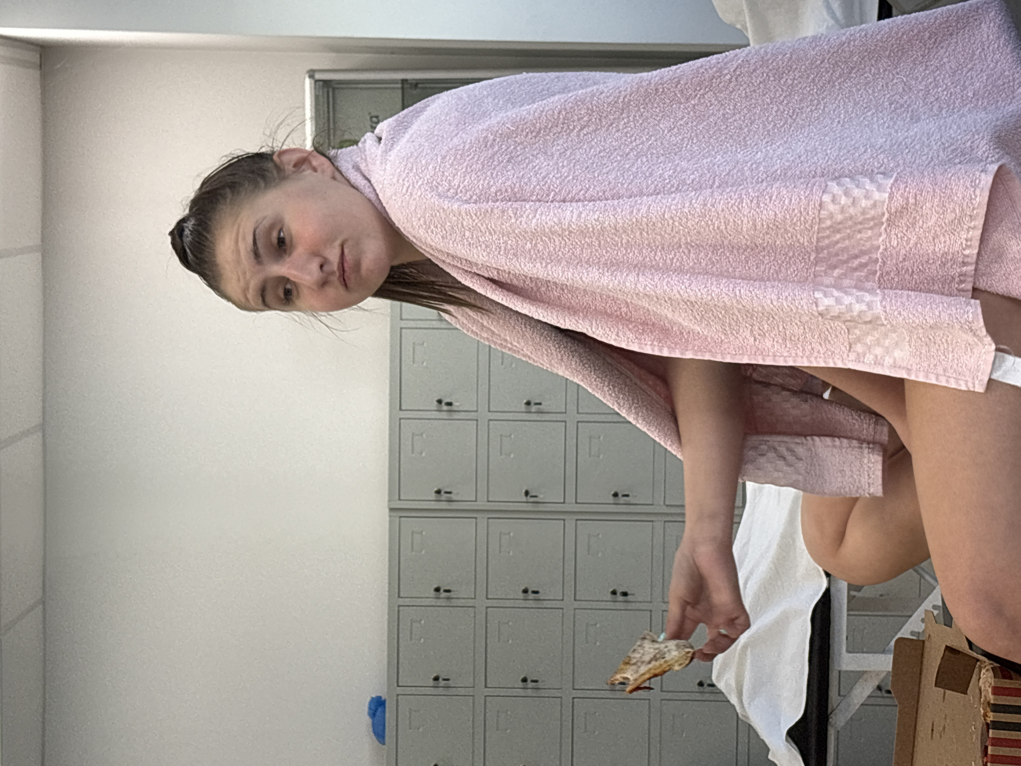 A woman in a pink towel stands in what appears to be a locker room. She holds a piece of food in her right hand. Behind her are rows of gray lockers. A white wall is visible to the left. A sink with a blue paper towel dispenser is attached to the wall. Parts of a white towel and a cardboard box are also visible. The scene is likely in a gym or recreational facility. The location is unknown, as there is nothing in the image that indicates a specific location in Sofia, Bulgaria.