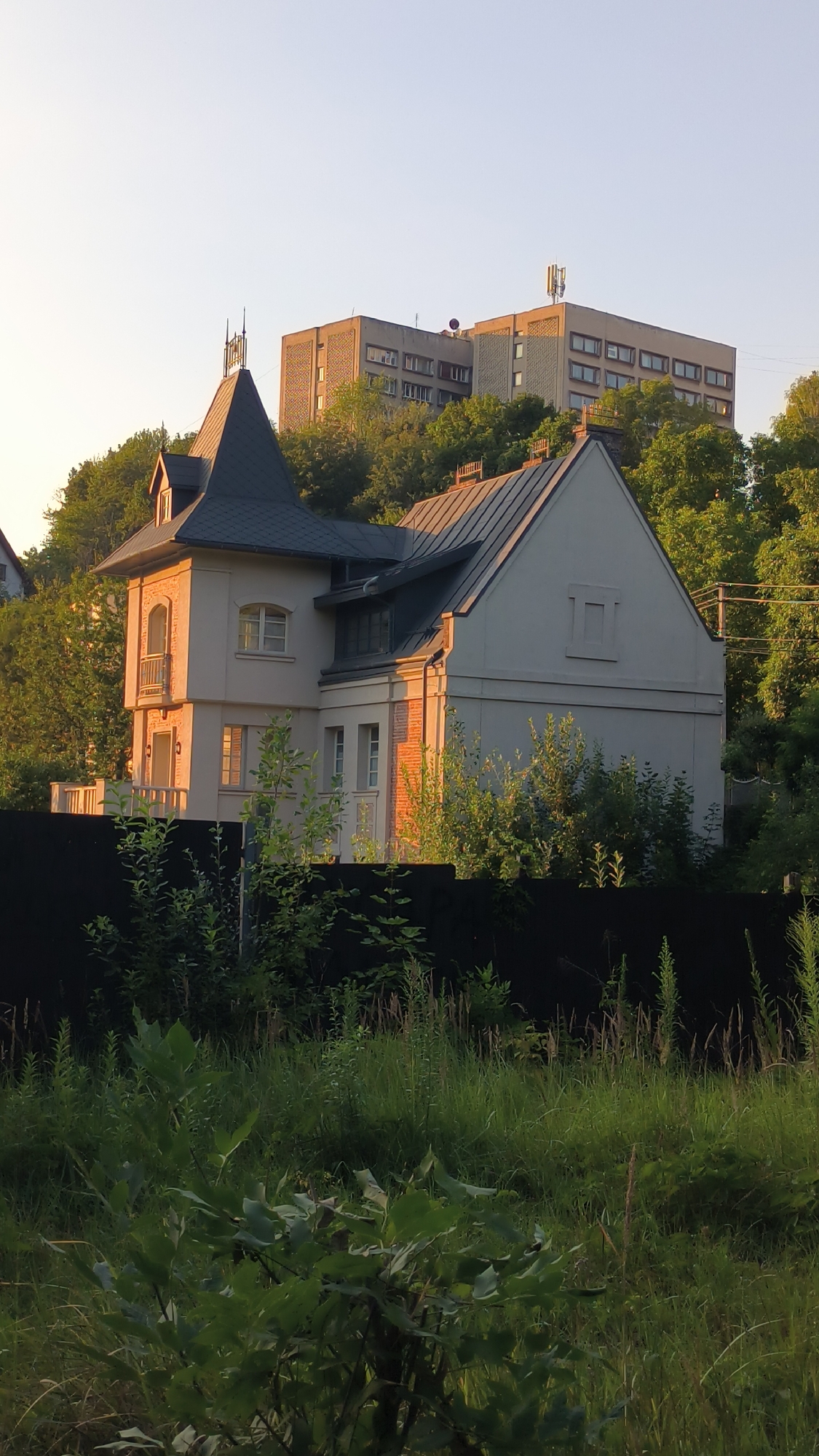 The image depicts a multi-story residential building situated on a hillside in Lviv, Ukraine. The building exhibits a modern architectural style with a light-colored facade and dark-colored roof. In the foreground, a dense growth of green vegetation, including trees and shrubs, partially obscures a black fence. The background features a larger, more utilitarian building, likely an apartment block, also set against a backdrop of lush green trees. The sky is clear and a pale yellow, suggesting either early morning or late afternoon sunlight. There are no people or vehicles visible in the scene. The overall impression is one of a residential area integrated with natural elements.