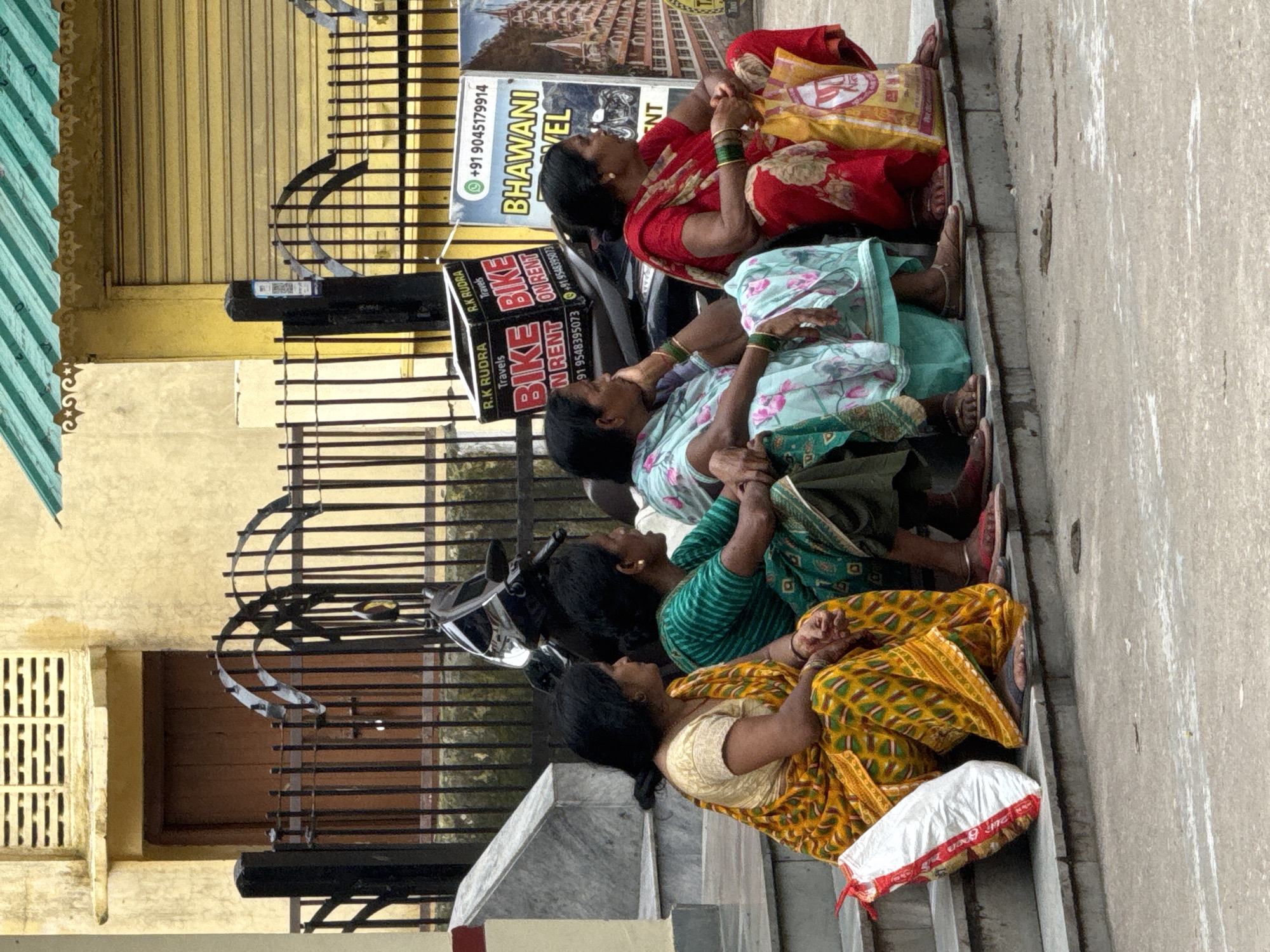 In Rishikesh, India, four women are seated on concrete steps outside a building with a yellow facade. The woman on the far left wears a yellow and green patterned sari. Next to her, a woman in a green and white striped sari is looking to her right. The third woman from the left wears a blue and pink floral patterned sari. The woman on the far right is dressed in a red sari and holds a yellow shopping bag with text. Behind the women, a black metal fence separates them from a scooter and posters. One poster advertises "R.K. RUDRA Travels BIKE ON RENT" with a phone number. Another poster features "+91 9045179914 BHAWANI TRAVEL". The environment appears to be an urban street in Rishikesh.