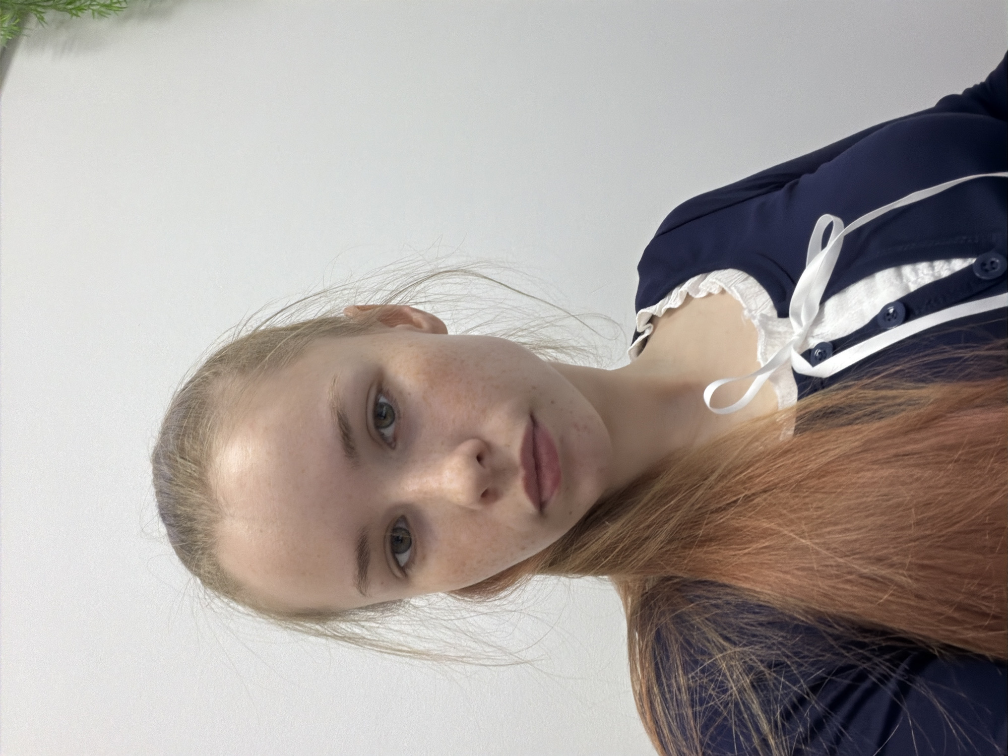 A young woman with fair skin and freckles is depicted in the image. She has light-colored hair and green eyes, wearing a dark blue top with white accents and a lace collar. The background is a plain, light gray/white. The image appears to be taken indoors. Location in Berezniki, Russia is not possible to determine.
