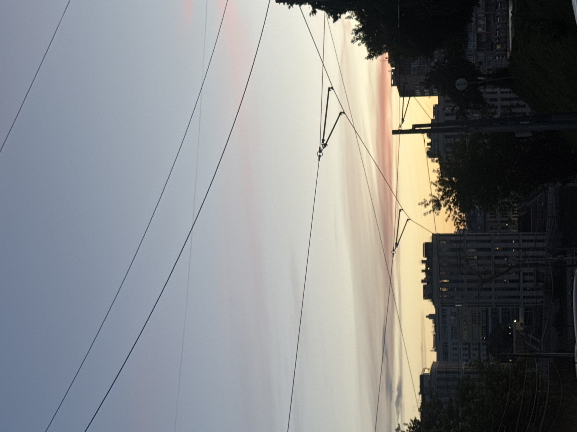 This image depicts a city skyline at dusk, with buildings silhouetted against a soft, pastel-colored sky. Thin wires crisscross the frame, suggesting an urban environment with overhead power lines or trolley lines. Several multi-story buildings are visible, varying in architectural style. In the foreground, trees and darker structures partially obscure the view, adding depth to the scene. The lighting suggests either early morning or late evening.
While the image shows an urban landscape, it is not possible to definitively identify the city as Nizhny Novgorod, Russia, based solely on the visual information provided. The style of architecture and the presence of overhead wires are common features in many cities worldwide. Therefore, a specific location within Nizhny Novgorod cannot be determined.