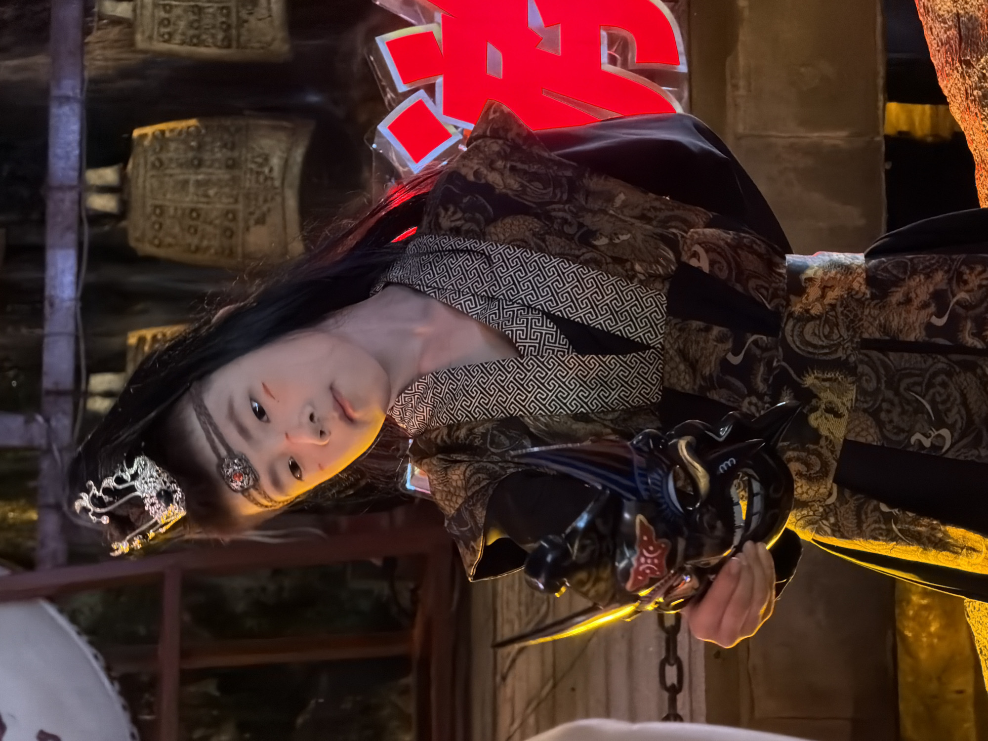 The image shows a young person wearing traditional Chinese attire, including a headdress and patterned robes. They are holding a decorative mask up to their face with their right hand. The person has theatrical makeup on their cheeks. In the background, large decorative bells are visible, and to the right, large red characters are illuminated. The setting appears to be an outdoor location at night, possibly part of a festival or cultural event. Based on available information, this scene is likely located in the Yuzhong District of China.