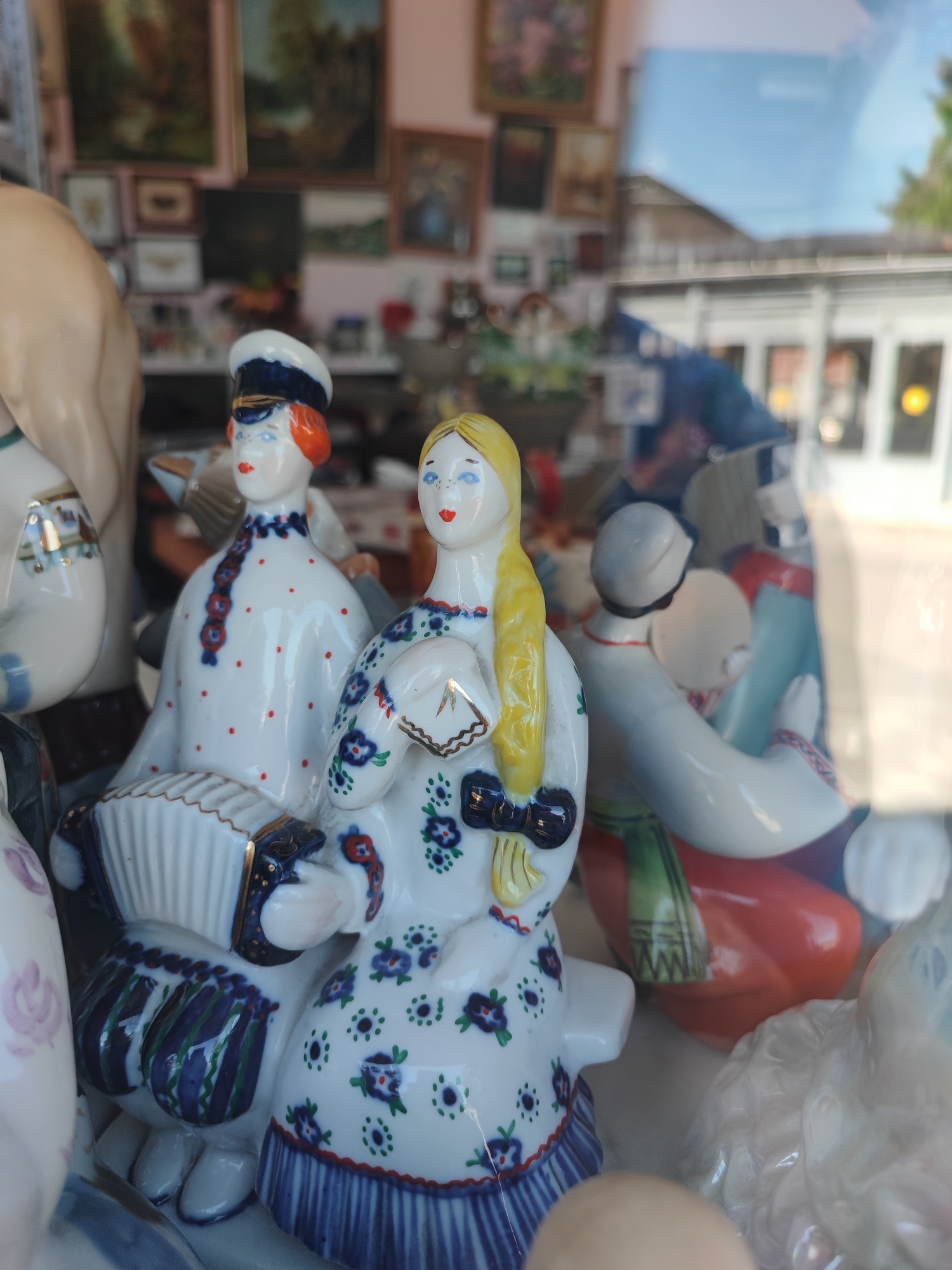 The image shows a collection of porcelain figurines displayed behind glass, likely in a shop window. The figurines depict people in traditional clothing, some holding musical instruments such as an accordion. The background is blurred, showing framed pictures on a wall and the reflection of a street scene with buildings and trees. The figurines are detailed with painted faces, colorful attire, and intricate patterns. This scene is located in Pyatigorsk, Russia, suggesting a possible souvenir shop or antique store.