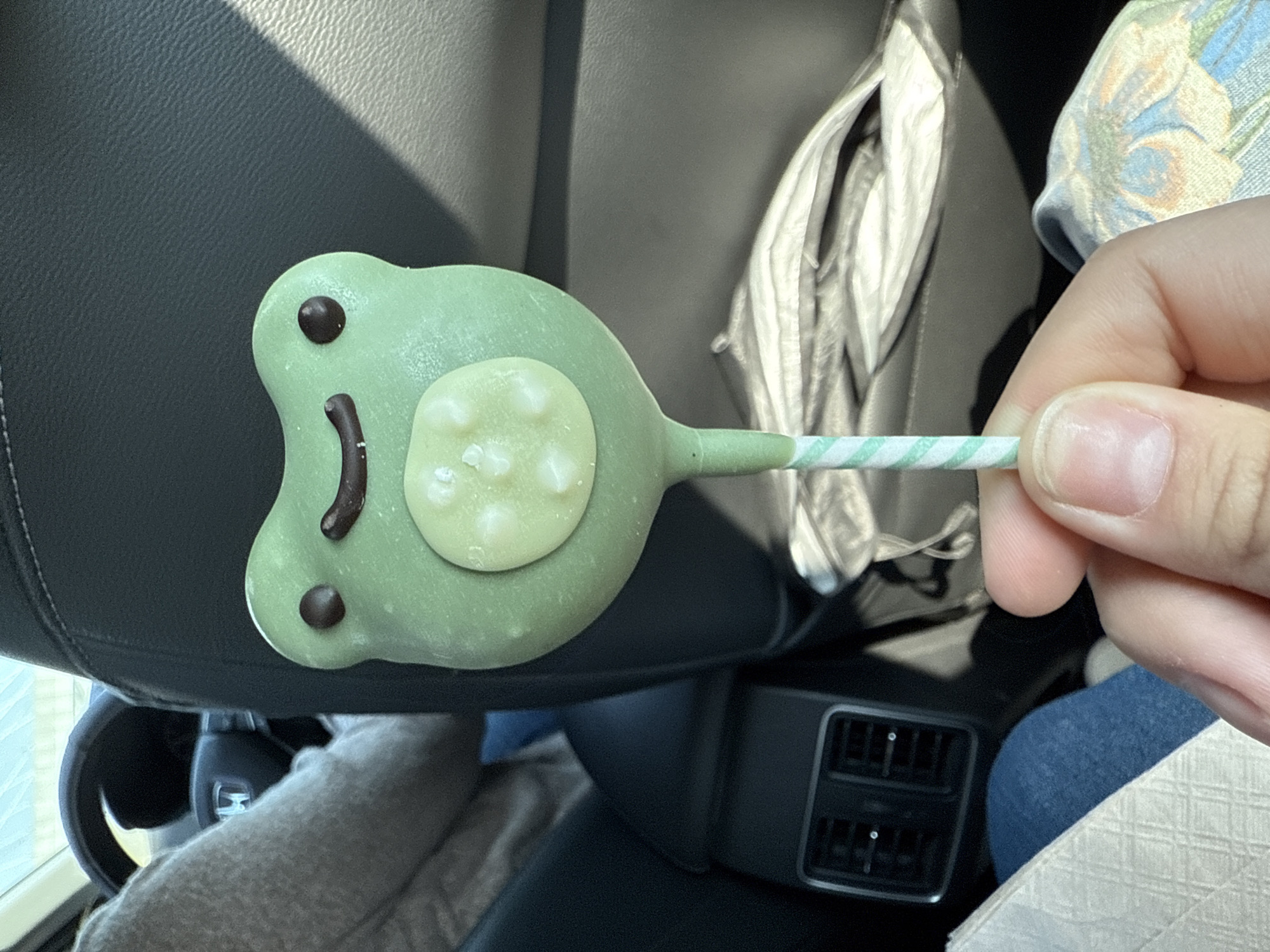 A hand holds a green frog-shaped lollipop with a green-and-white striped stick. The lollipop has brown eyes and a brown mouth. A light-colored circle with white spots is on the frog's body. The background includes a car seat, the back of the front seat, and a blue-jeaned leg.