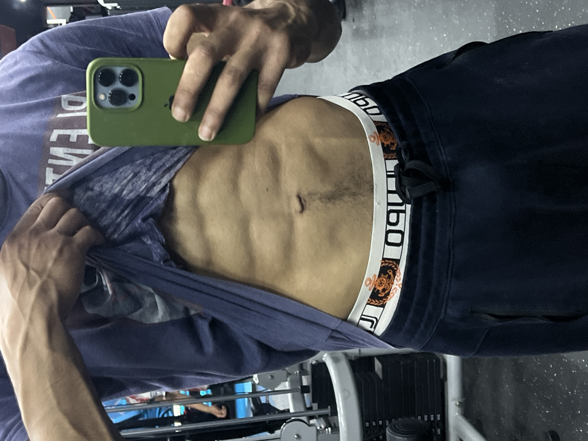 A person in a gym setting takes a selfie in M'Diq, Morocco, partially lifting their shirt to display their abdominal muscles. They are holding a green iPhone, and their navy-colored sweatpants are visible below. Gym equipment is visible in the background.