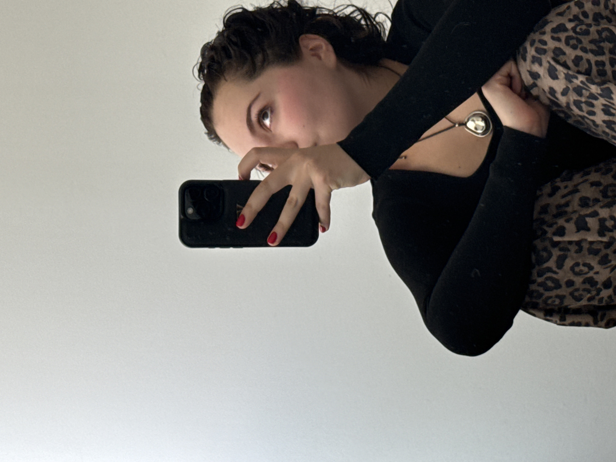 A woman with curly dark hair is holding a black smartphone in her left hand, taking a selfie. Her fingernails are painted red. She is wearing a black long-sleeved shirt and animal print pants or leggings. She has a silver pendant necklace. The background is a plain white wall. The location appears to be indoors, but there is no clear evidence to determine if it is in Mexico City.