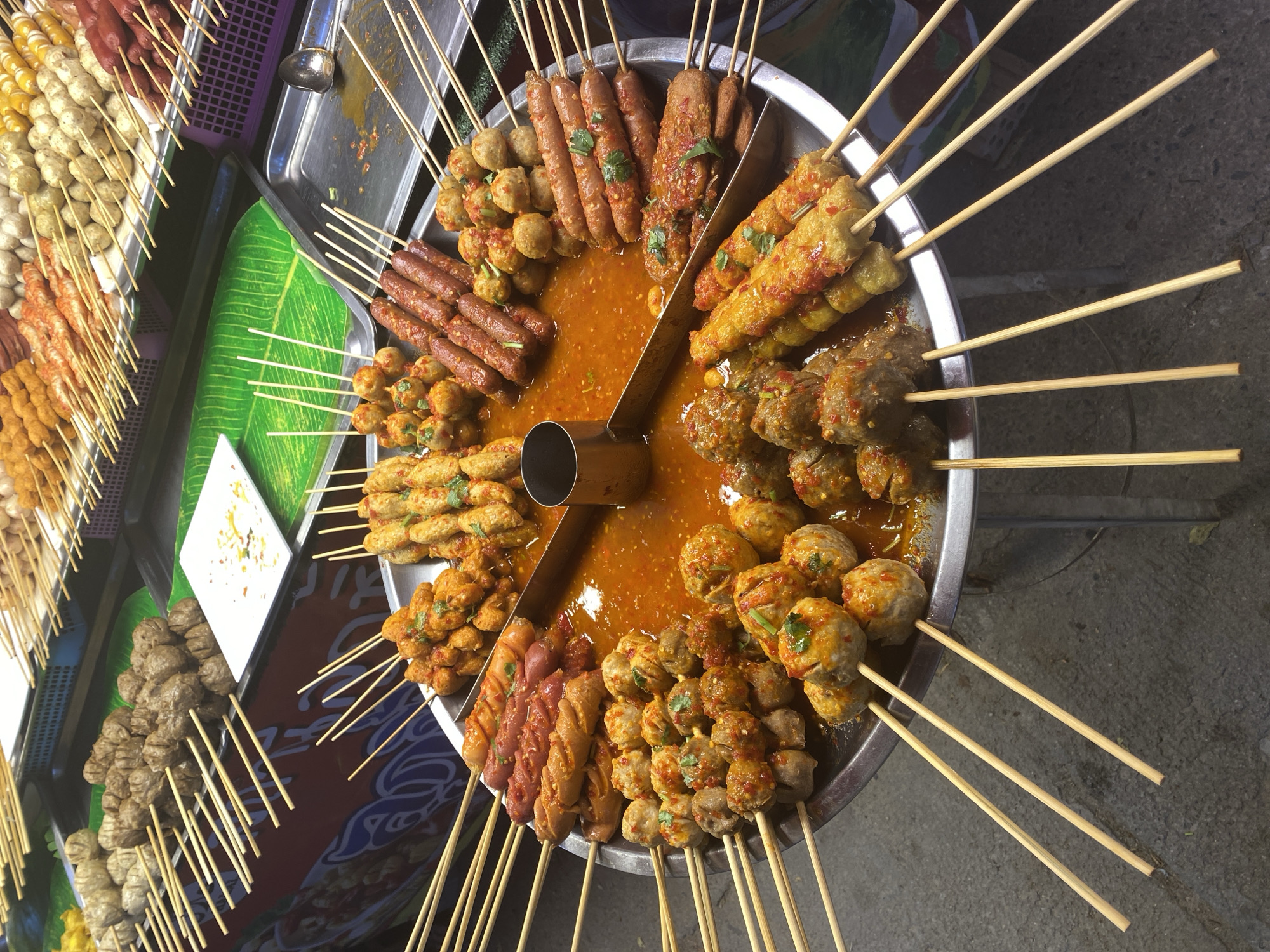 A street food vendor in Baan Mae Nam, Thailand, displays a variety of grilled skewers, primarily meatballs and sausages, arranged radially around a central container of dipping sauce on a large metal tray. The skewers are displayed on a metal tray. Adjacent to the main display are additional trays of skewers, including rows of meatballs of varying colors and coatings. A green banana leaf lies flat on one of the trays. The scene is lit by artificial light. The surface beneath the display is concrete.