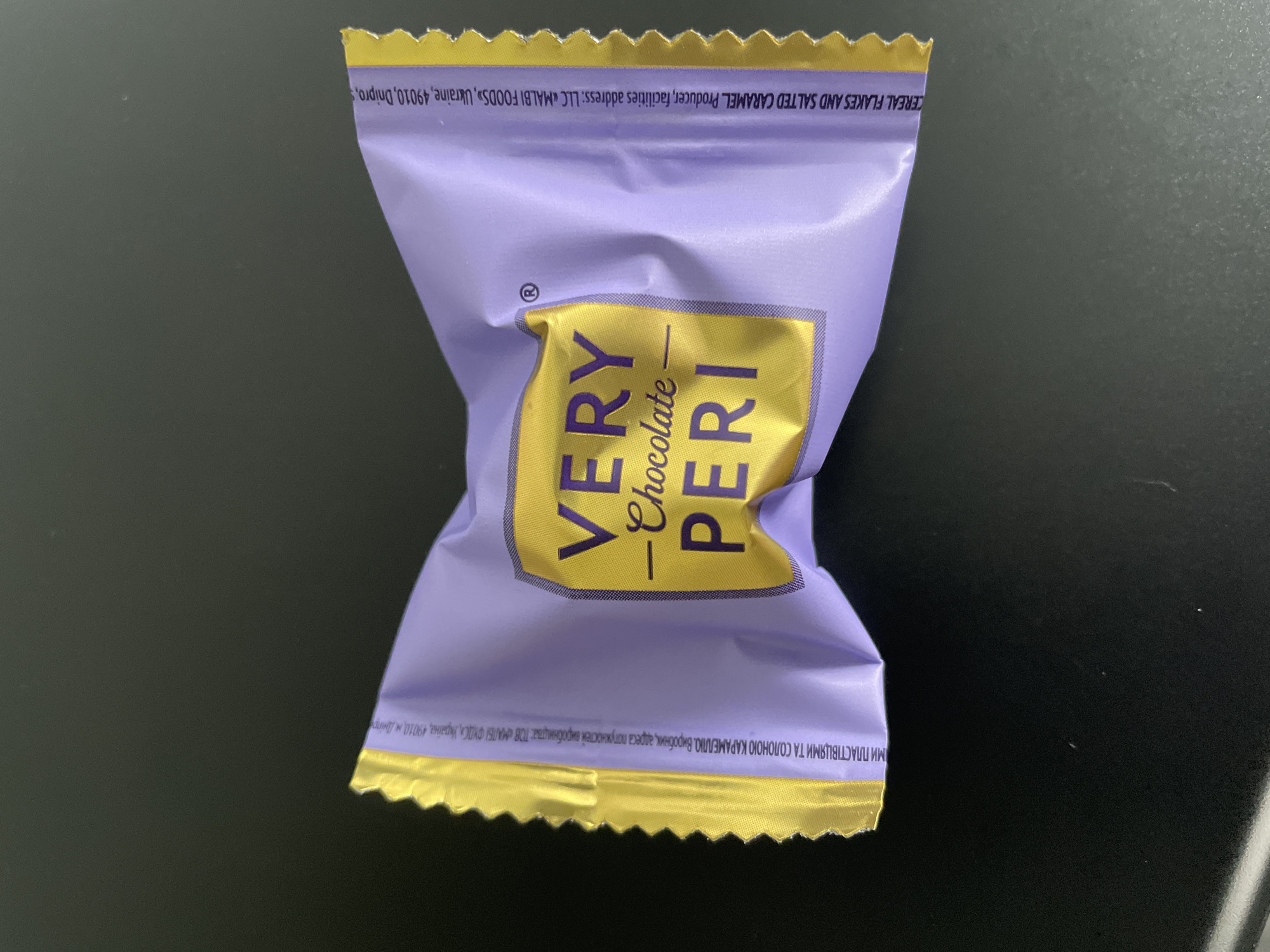 A close-up shot of a purple-and-gold wrapped candy called "Very Chocolate Peri." The wrapper indicates it contains cereal flakes and salted caramel. The wrapper also lists the producer as "LLC MALBI FOODS" in Dnipro, Ukraine. The candy is on a dark surface.