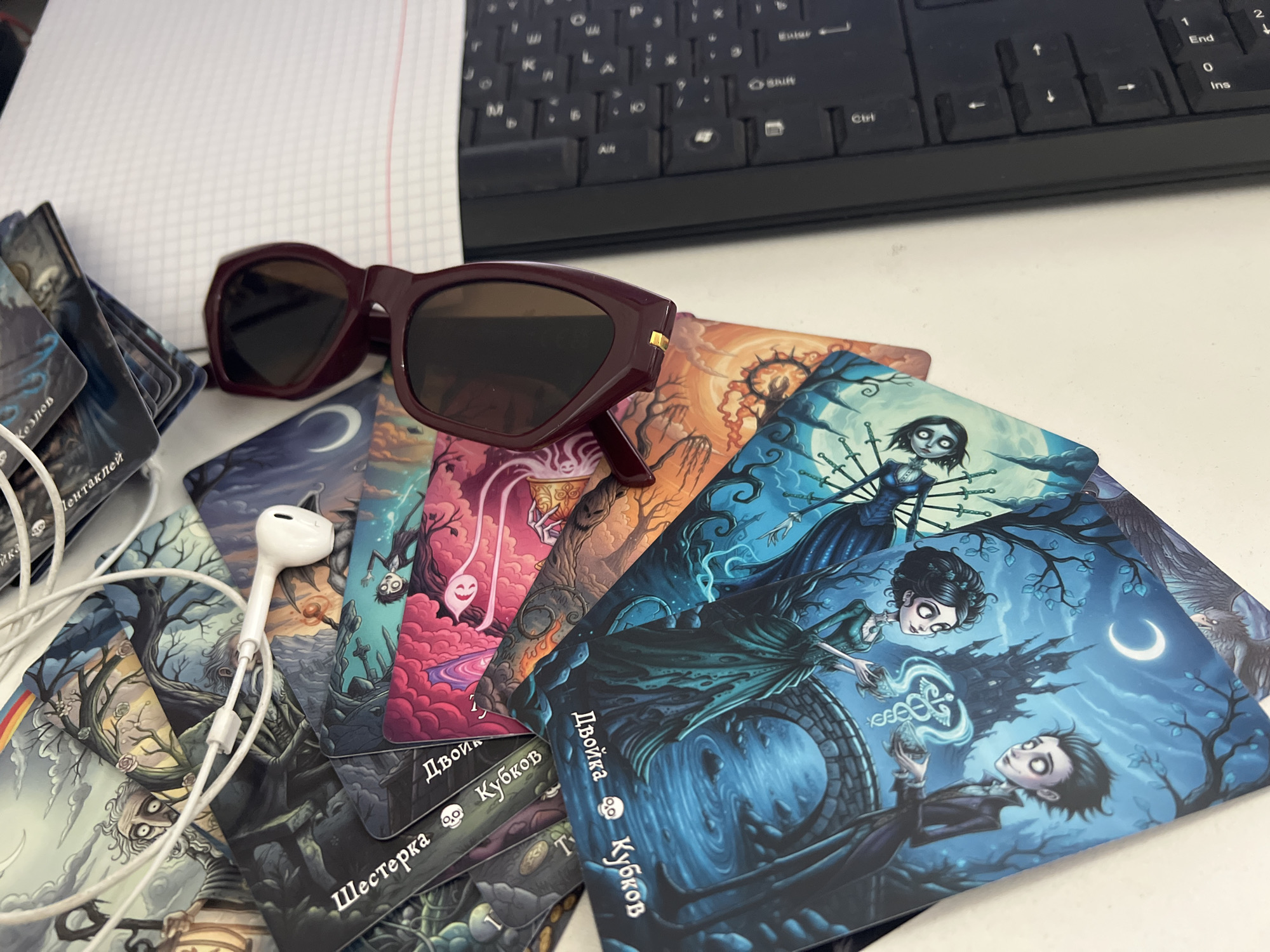 The image shows a close-up, top-down view of a white desk surface. On the desk are several items, including a stack of tarot-like cards with gothic-themed illustrations, a pair of dark red sunglasses, and a white wired earbud. A portion of a laptop keyboard is visible in the upper right corner, and a sheet of graph paper is in the upper left. The cards are fanned out, with the topmost cards featuring illustrations of skeletal figures and dark, whimsical landscapes. The sunglasses are positioned on top of the cards. The earbud is lying on top of some of the cards, with its white wire extending across them. The overall lighting suggests an indoor setting. There is no specific visual information in the image that would allow for the identification of a location in Almaty, Kazakhstan.