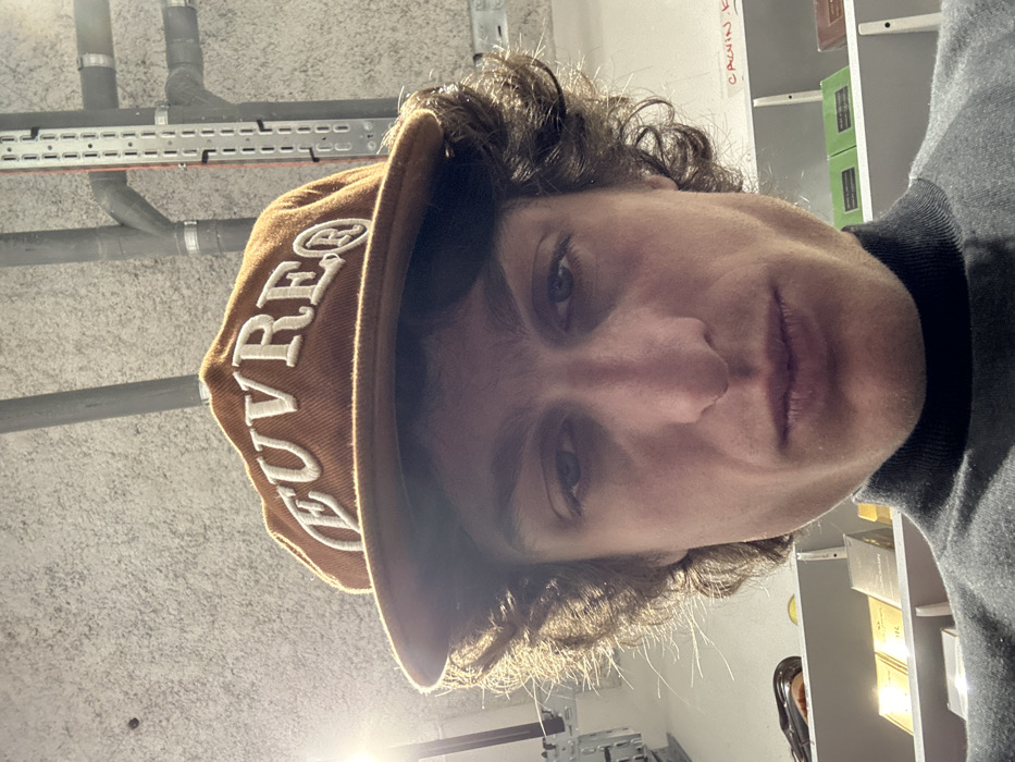 An individual is depicted in a close-up, low-angle indoor shot, positioned in Joinville-le-Pont, France. The male subject has fair skin, dark curly hair, and wears a brown baseball cap with "EUVRE®" embroidered in white capital letters on the front. He is also wearing a dark grey turtleneck sweater. The subject looks directly at the camera with a neutral expression. The background features an industrial-style ceiling with exposed pipes and a perforated metal cable tray. Below this, white shelving units are visible, displaying numerous packaged items, including various small, rectangular boxes in metallic and green finishes, suggesting products like fragrances or cosmetics. A handwritten label, partially obscured on a shelf to the right, appears to read "CALVIN K". A bright light source is present in the upper left portion of the image.