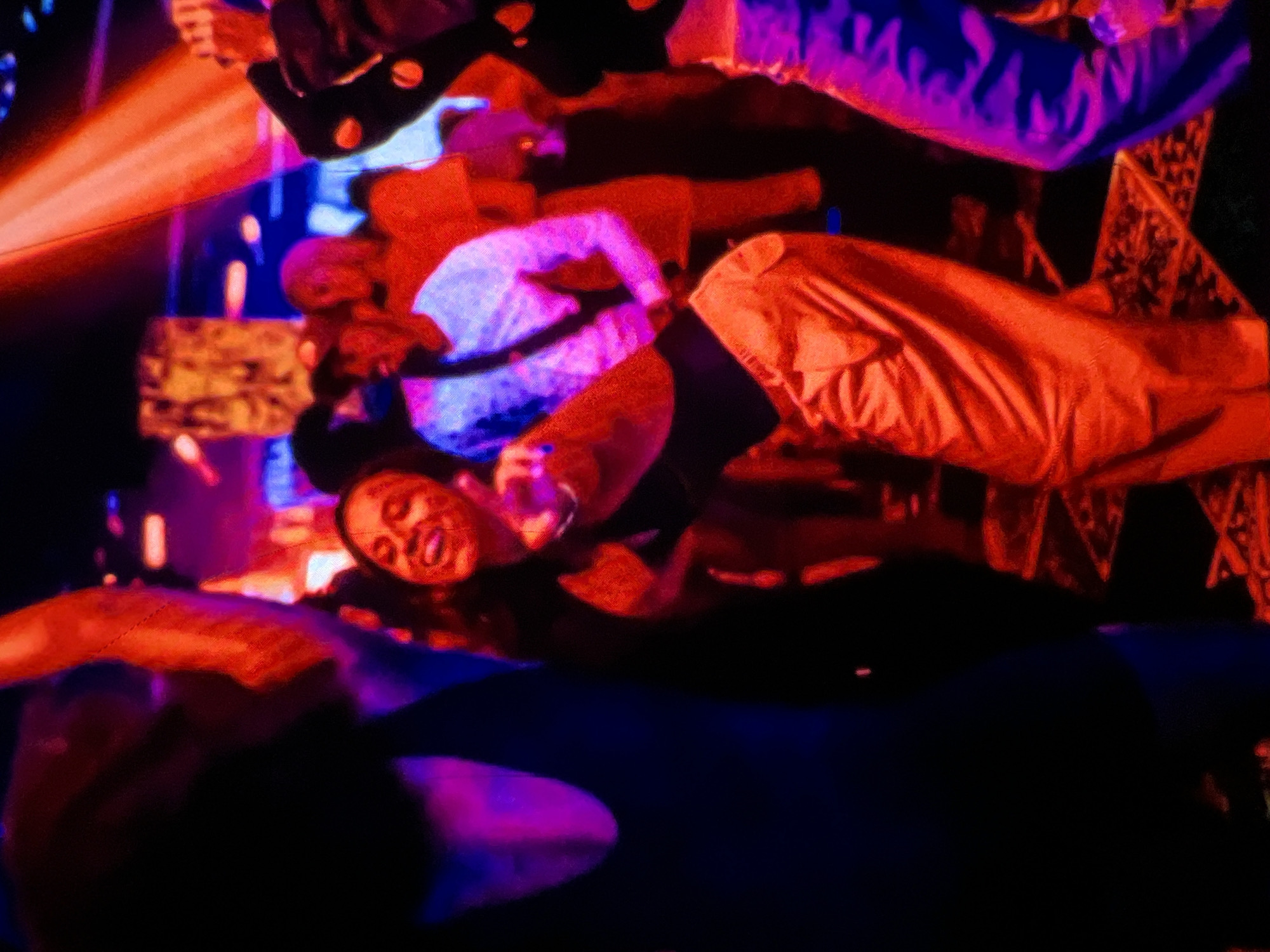 A group of individuals are dancing in a dimly lit, vibrant environment, likely a nightclub or party venue in Lagos, Nigeria. The scene is dominated by red and blue lighting, creating a dynamic and energetic atmosphere. In the foreground, a woman with dark hair is enthusiastically dancing, her face illuminated by the red light, and she is captured mid-gesture with her arm raised and finger pointing. She is wearing an orange outfit. To her left, another person, mostly obscured by darkness, appears to be dancing as well, with a portion of their dark clothing visible. In the background, several other people are present, their forms softened by the low light and movement. One individual in the mid-ground is wearing a light-colored shirt and dark pants, and another person to their right is wearing a dark jacket with visible circular embellishments and light-colored pants. The floor has a patterned design, suggesting a deliberate aesthetic for the venue. The overall impression is one of a lively social gathering and celebration.