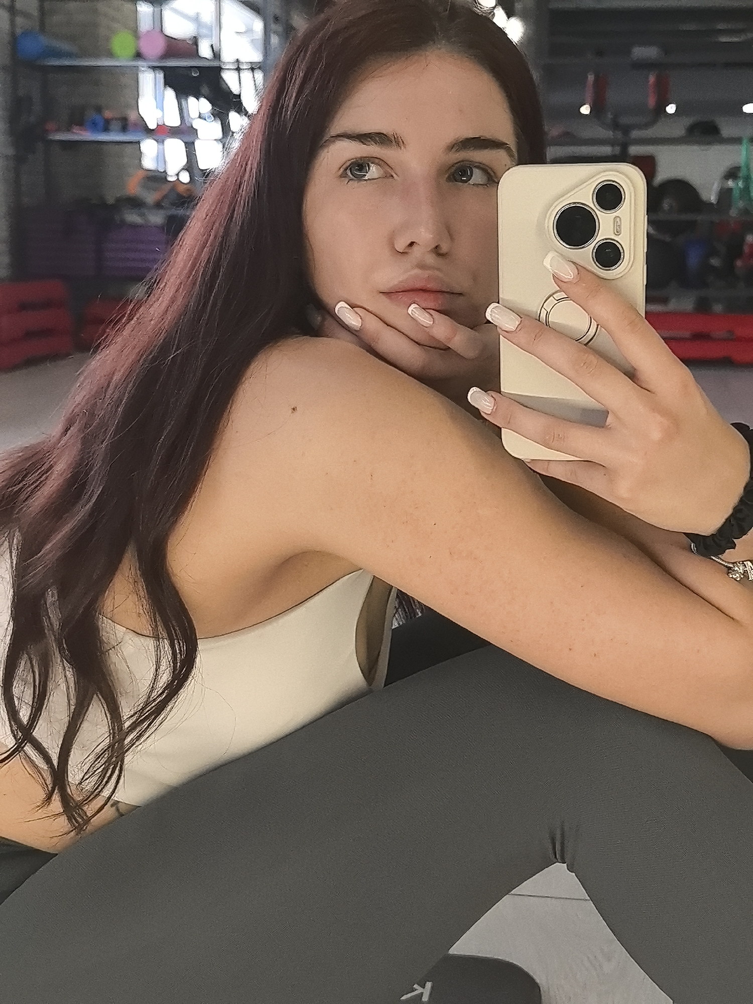 A young woman with long dark brown hair and light skin is taking a selfie in a gym. She is wearing a white sports bra and dark gray leggings. Her left arm is resting on her leg, and her right hand is holding a white smartphone, which is capturing the image. The background shows gym equipment, including shelves with weights and mats. The setting appears to be an indoor fitness facility.
