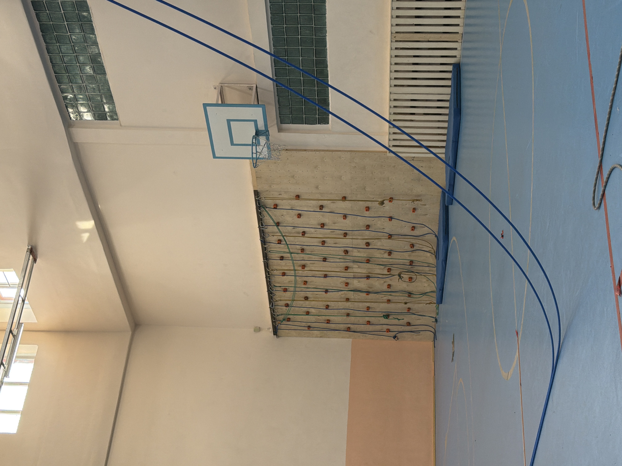 The image shows the interior of a gymnasium in Berezniki, Russia. The gym features a blue floor with white and red lines marking the court, and a wall-mounted rock climbing structure with red climbing holds and associated safety ropes. Above the structure is a basketball hoop. The walls are painted in neutral tones, with a section of pink near the climbing wall. There are windows on the left side of the image. Above the basketball hoop, there are glass block windows and slatted ventilation. A blue rope is hanging across the space.