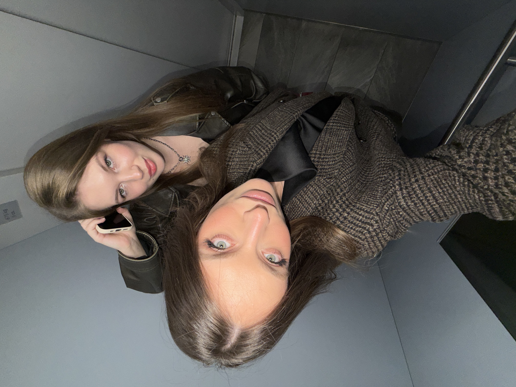 Two women are in an elevator, posing for a selfie-style photograph. One woman is on the phone, wearing a leather jacket and a necklace with a sun pendant. The other woman is wearing a patterned brown coat over a black top. The elevator has gray walls and flooring, with a silver handrail. The location is Vozdovac Urban Municipality, Serbia.