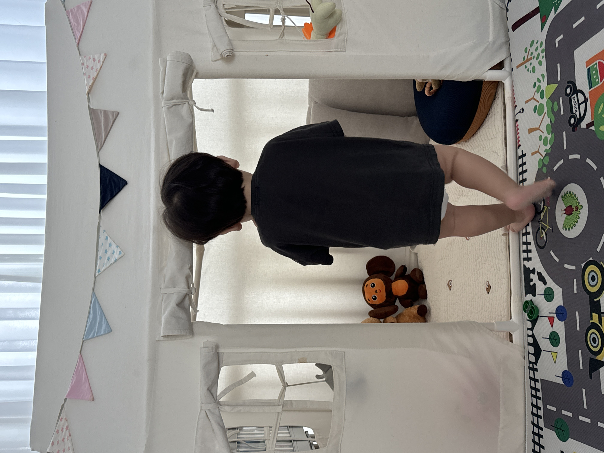 A young child stands with their back to the camera, facing away from the viewer and towards the interior of a white canvas play tent. The child is wearing a dark t-shirt and appears to be wearing a diaper. The tent has decorative triangular flags strung across its top, featuring various patterns including pink with white stars, solid pink, light blue with white stars, and solid dark blue. Two windows are visible on the left side of the tent, appearing to be covered by fabric flaps tied back. Inside the tent, a plush toy monkey with large ears and an orange face is visible on the floor, along with another brown plush toy to its left. The floor inside the tent is covered with a light-colored rug with a subtle pattern. To the right of the child, a dark blue cushion with an orange base is visible. Outside the tent, a play mat with a road and cartoon vehicle design is on the floor. The background consists of white vertical curtains. This scene is likely in a home environment within Jeju, South Korea, given the context provided.