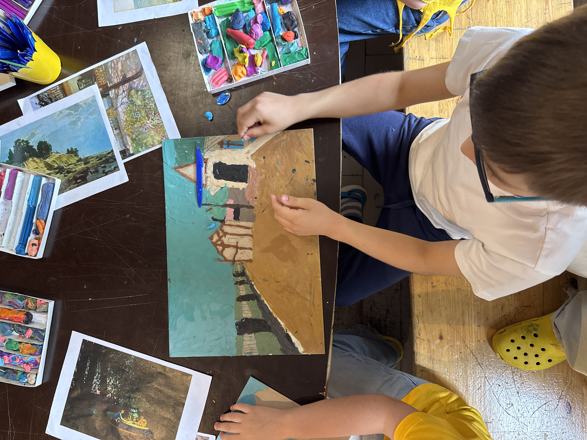 This is an overhead, top-down view of a table where children are engaged in a crafting activity, likely involving clay or plasticine. The scene appears to be an indoor setting, possibly a classroom or a home art studio, with a dark brown, scratched table surface and a lighter wooden table beneath.
Several children's hands are visible, actively working on or near the crafting materials. One child, wearing a yellow t-shirt and blue jeans, has their hands positioned over a piece of artwork created with what appears to be clay. This artwork depicts a landscape with a house and a fence. Another child, wearing a white t-shirt and blue pants, is also working with clay, placing small pieces onto the artwork. A third child, partially visible on the right, is also present with their hands near the crafting supplies.
Scattered across the table are various art supplies, including boxes of colored clay or plasticine, individual pieces of clay, and some printed reference images. One box contains a colorful assortment of clay sticks. Another box holds individual lumps of various colored clay. A yellow cup filled with blue drawing utensils is also visible.
The children are seated, and their legs and feet, clad in jeans, yellow Crocs, and blue pants, are visible at the bottom of the frame. The overall impression is one of focused creative activity.
This scene is not located in Moscow, Russia. The provided image does not contain any information or visual cues that would indicate a specific location, let alone Moscow.