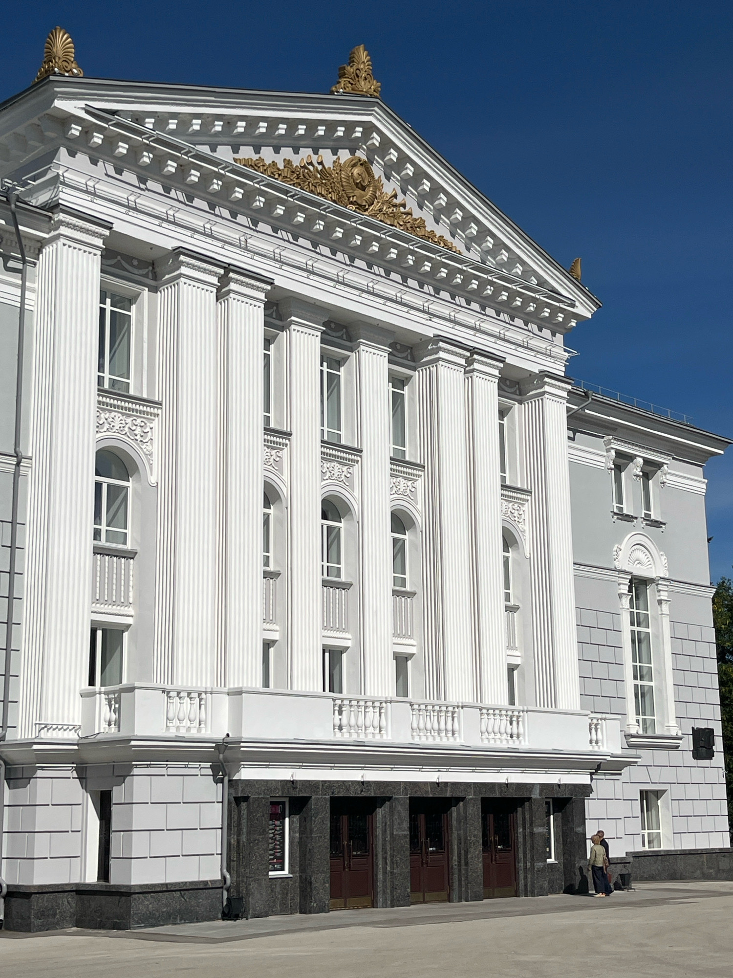 The image shows the Perm Academic Theatre-Theatre on a sunny day. The theatre is a large, classical building with a white facade adorned with fluted columns, arched windows, and ornate gold detailing on the pediment. A balcony with a balustrade extends across the upper level. The lower level features dark granite cladding and several large double doors. Two individuals, a woman and a man, are standing near the entrance. The sky is a clear, vibrant blue.