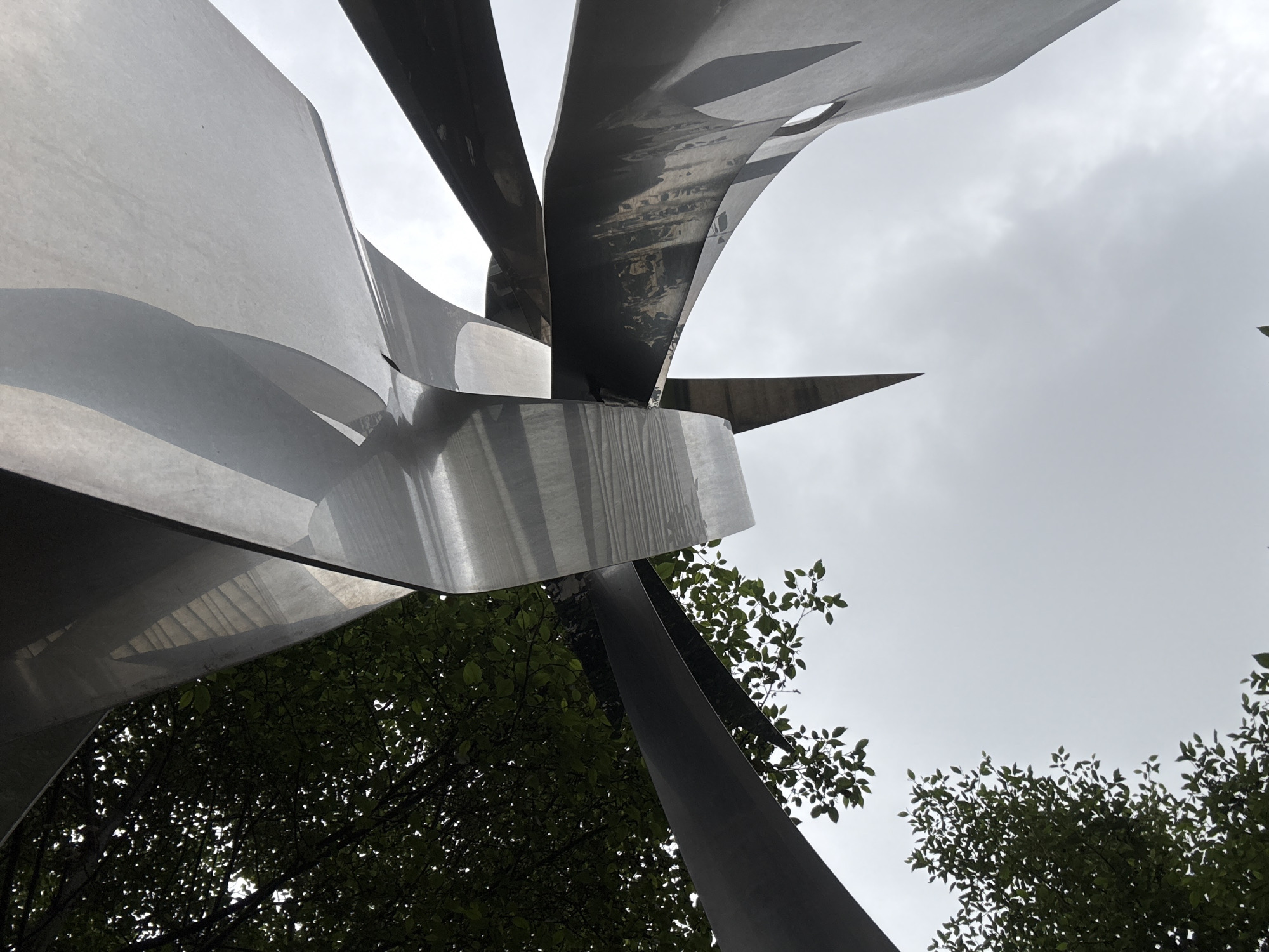 A low-angle shot of a stainless steel sculpture under a cloudy sky, with tree foliage visible at the base of the sculpture. The sculpture features multiple curved, overlapping metal plates and angled segments extending outward from a central point. The location is likely an outdoor area in Mexico City, Mexico, based on image content and metadata. No people are visible in the image.