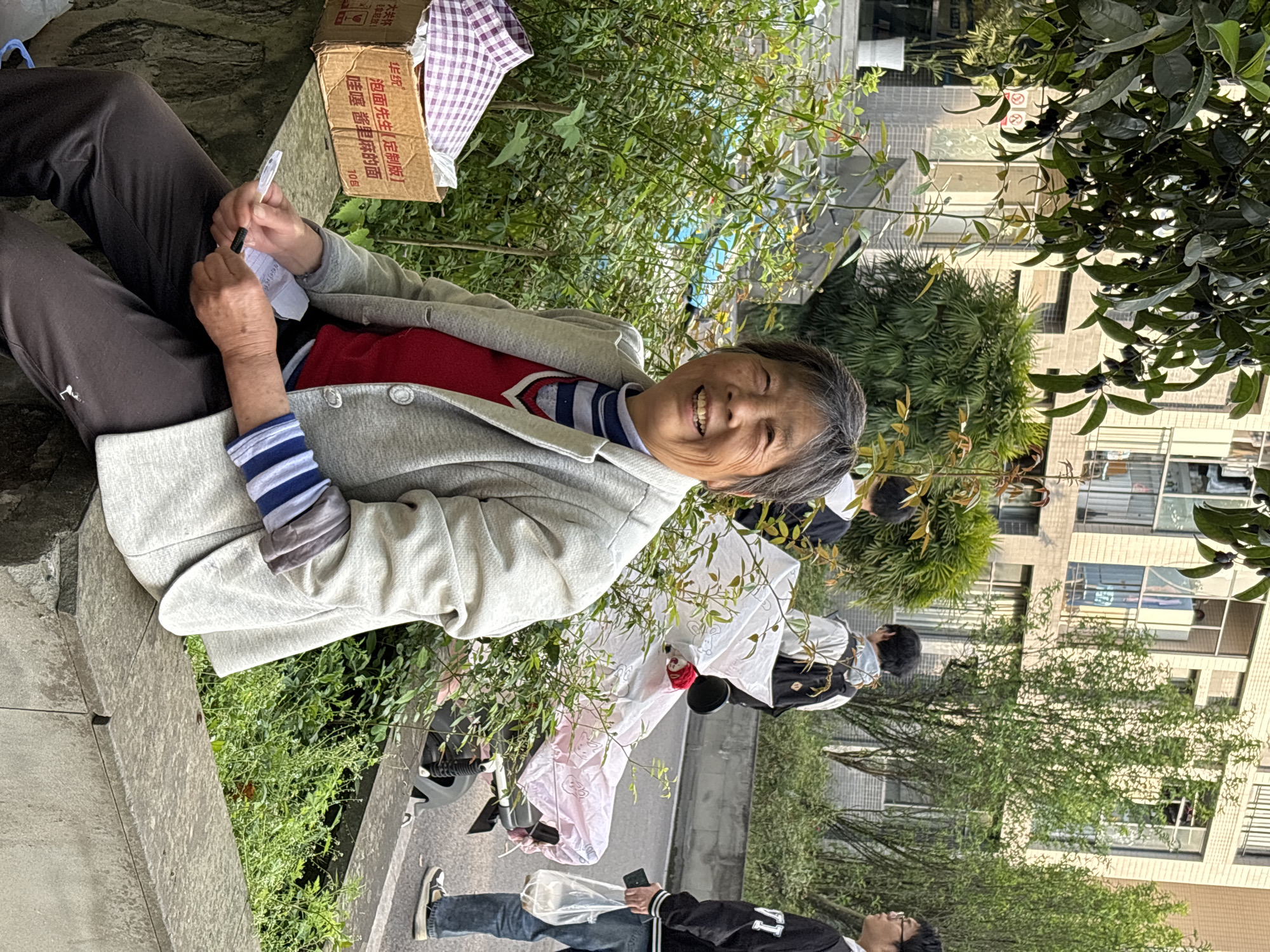 The image shows an elderly Asian woman seated outdoors in Jiangjin, China. She is positioned near a low concrete wall overgrown with greenery. The woman is writing on a piece of paper using a pen. Next to her is a cardboard box labeled with Chinese characters. She is smiling at the camera.
In the background, there are people walking on a sidewalk. Further back, there's a multi-story building with windows visible behind trees and foliage.