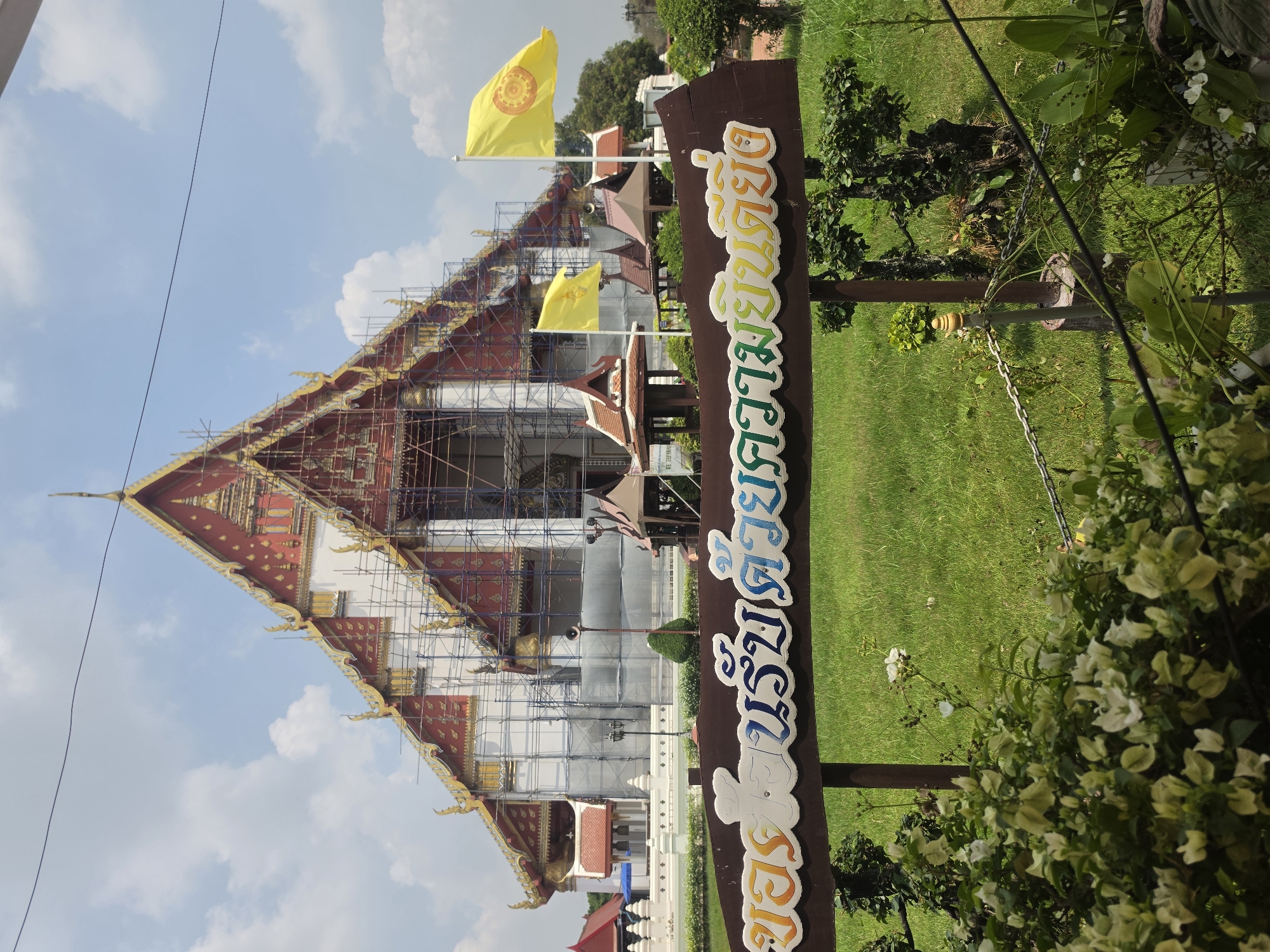 The image shows a temple in Phra Nakhon Si Ayutthaya City Municipality, Thailand. Scaffolding covers much of the building, suggesting it is under renovation. Two yellow flags fly near the temple. A wooden sign with colorful Thai script stands in the foreground, partially obscured by foliage. Green grass and trees surround the temple. The sky is blue with scattered clouds.