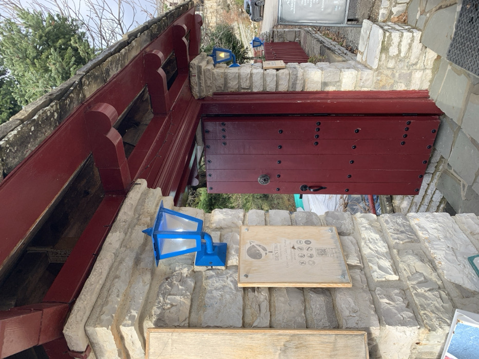 This image captures a detail-rich entrance to a building in a mountainous village, typical of Vikos, Greece. The scene is static, devoid of people, and appears to be daytime under an overcast sky, with bare trees suggesting late autumn or winter.
The central feature is a robust, dark red wooden door, constructed from vertical planks reinforced with numerous black studs arranged in rows. It is fitted with an ornate, circular metal handle/knob and a simpler dark handle below. The door is set into a wall of rough-hewn, light-colored stone blocks.
Above the entrance, a distinctive dark red wooden overhang or balcony extends, supported by decorative wooden brackets that resemble stylized figures. Two striking blue, angular electric lanterns are mounted on the stone walls, one prominently in the foreground and another further back near the door; both are illuminated with a warm light.
A light-colored wooden sign is attached to the stone wall in the foreground. It features a circular logo (possibly a stylized bird or plant) and the word "DENTIRO" (or similar) in capital letters. Below this, smaller text and a grid of icons are visible, including symbols for Wi-Fi, no smoking, and food, along with a QR code.
In the background, a steep, tree-covered hillside rises. A paved road is visible, with a grey hatchback car parked on it. Another larger, dark sign stands further up the slope, prominently displaying "VÍKOS" in large white letters. Additional Greek text below it, such as "ΕΝΟΙΚΙΑΖΟΜΕΝΑ ΔΩΜΑΤΙΑ" (Rooms to let) and "ΠΑΡΑΔΟΣΙΑΚΑ ΠΡΟΪΟΝΤΑ" (Traditional products), suggests it's an establishment offering accommodation and local goods. The overall impression is one of traditional architecture in a rugged, natural setting.