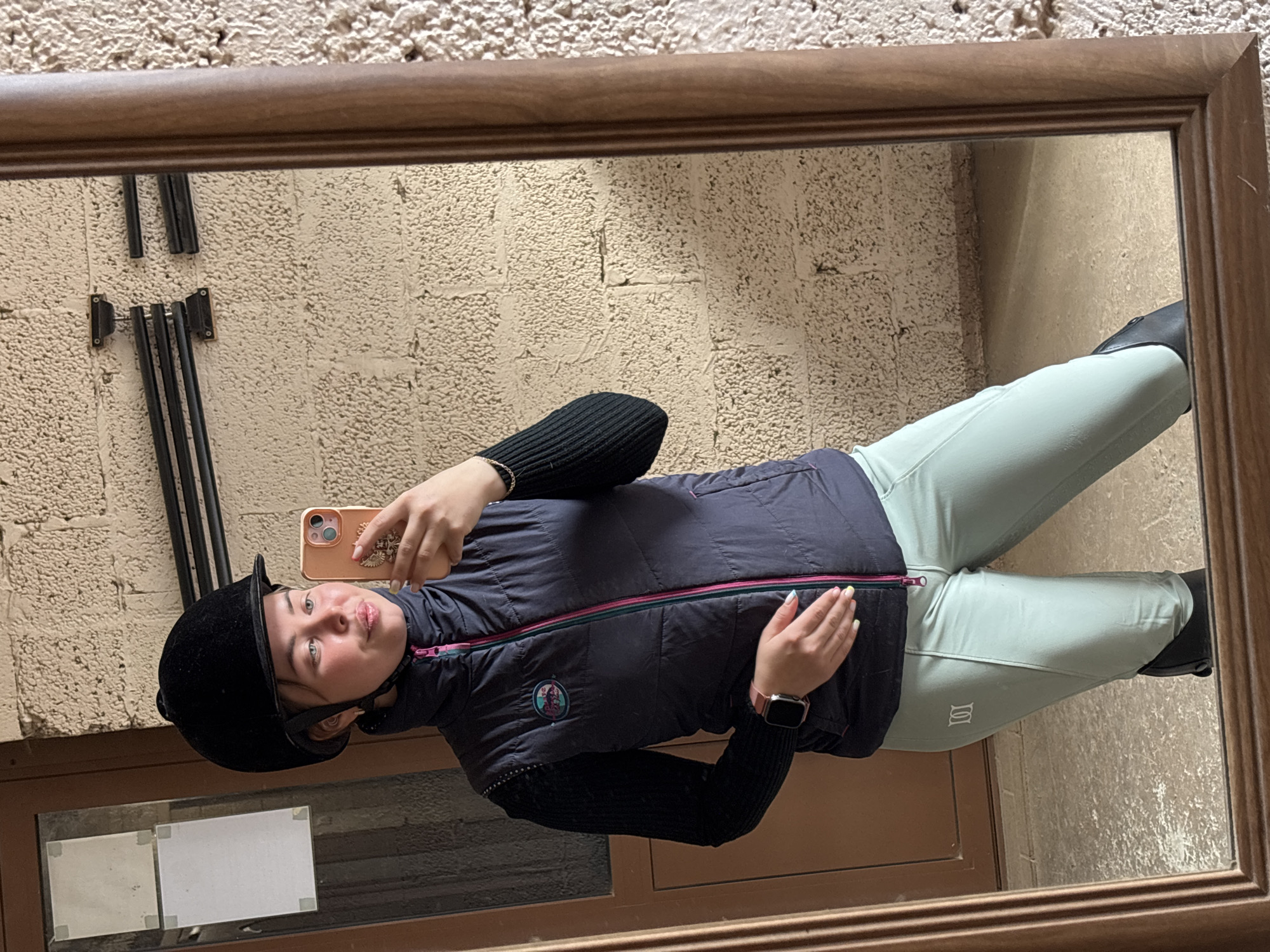 A young person, likely female, takes a mirror selfie while dressed in equestrian gear. She is wearing a black velvet riding helmet, a dark grey or black puffer vest with a bright pink zipper, a black long-sleeved top, and light mint green riding breeches. Her left wrist displays a smartwatch, and she holds a peach-colored smartphone with her right hand, which has light yellow/green painted fingernails. Black riding boots are visible on her feet.
The person is reflected in a large, wooden-framed mirror. The background, visible in the mirror, shows a textured, light-colored wall, possibly stone or rough plaster, with several black metal fixtures, appearing to be coat hooks or a rack. Below the mirror, a dark brown surface, possibly a door or floor, is visible. The image appears to be taken indoors, likely at an equestrian facility or home, in Русский Вожой, Russia. A white, stylized logo resembling two intertwined 'S' shapes is visible on the left thigh of her breeches.