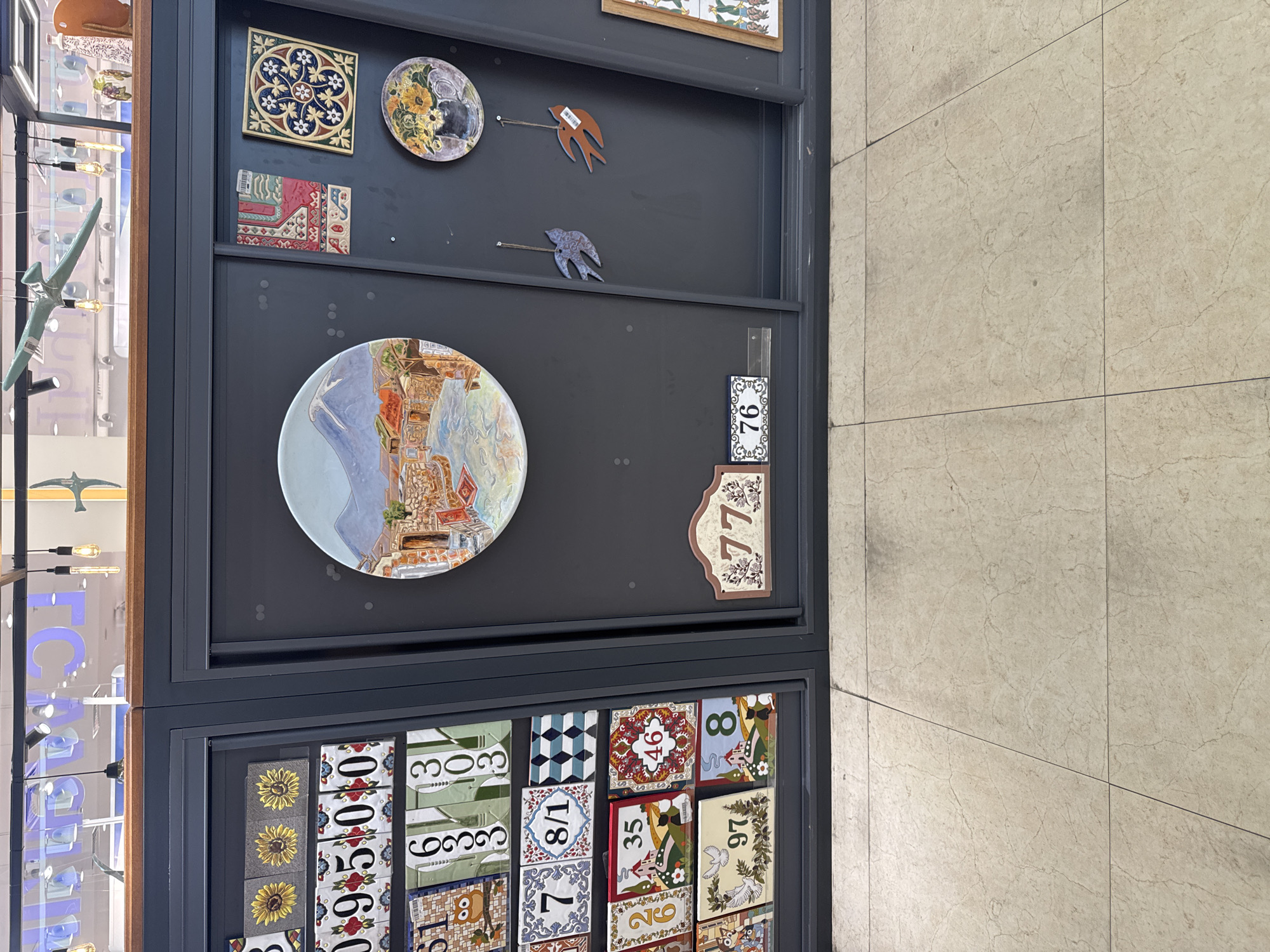 The image depicts a display shelf in what appears to be a retail environment. The shelf is dark gray and contains various decorative ceramic tiles and plates. On the left side of the shelf, there are rows of numbered tiles, some featuring patterns like sunflowers, flowers, and geometric designs. There are also tiles with house and animal motifs. Further right, a large circular plate is displayed, depicting a landscape with a mountain, buildings, and a body of water. To the right of this plate are smaller decorative tiles and two bird-shaped ornaments hanging from strings. Below the circular plate, there are two more numbered ceramic signs, "77" and "76". The floor in front of the display is tiled with light-colored square tiles. The overall environment suggests a shop selling home decor items.