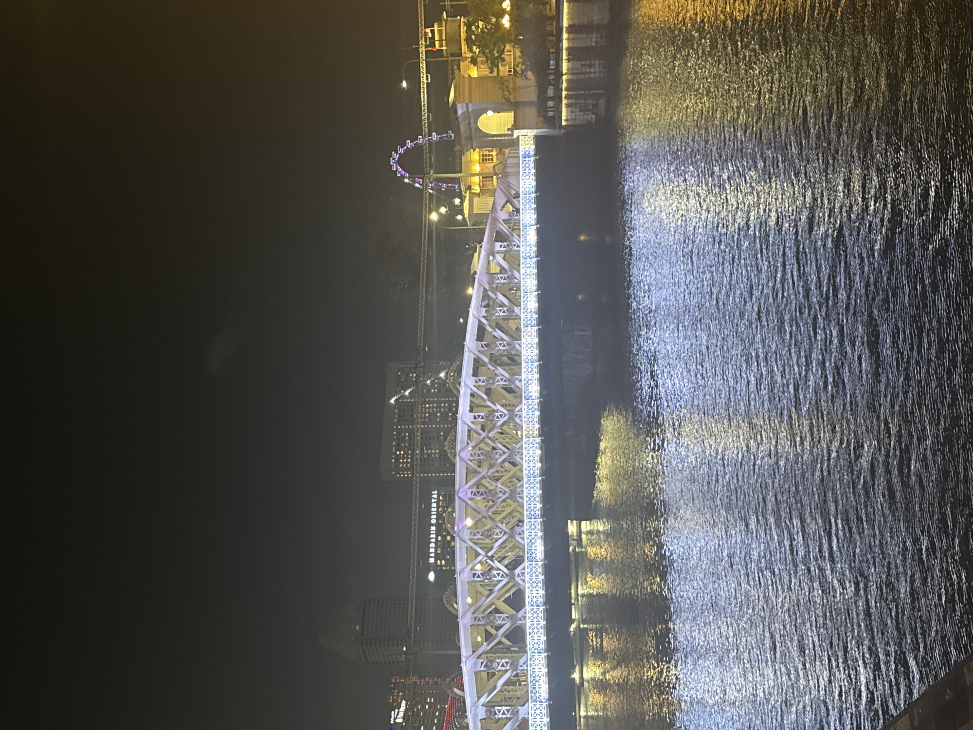 This is a night time, medium-angle, outdoor shot of a bridge spanning a body of water. The bridge is illuminated by white and purple lights. In the background, there are buildings, a Ferris wheel, and a sign that reads "Mandarin Oriental". The water reflects the lights from the bridge and the buildings. The overall scene is a cityscape at night.