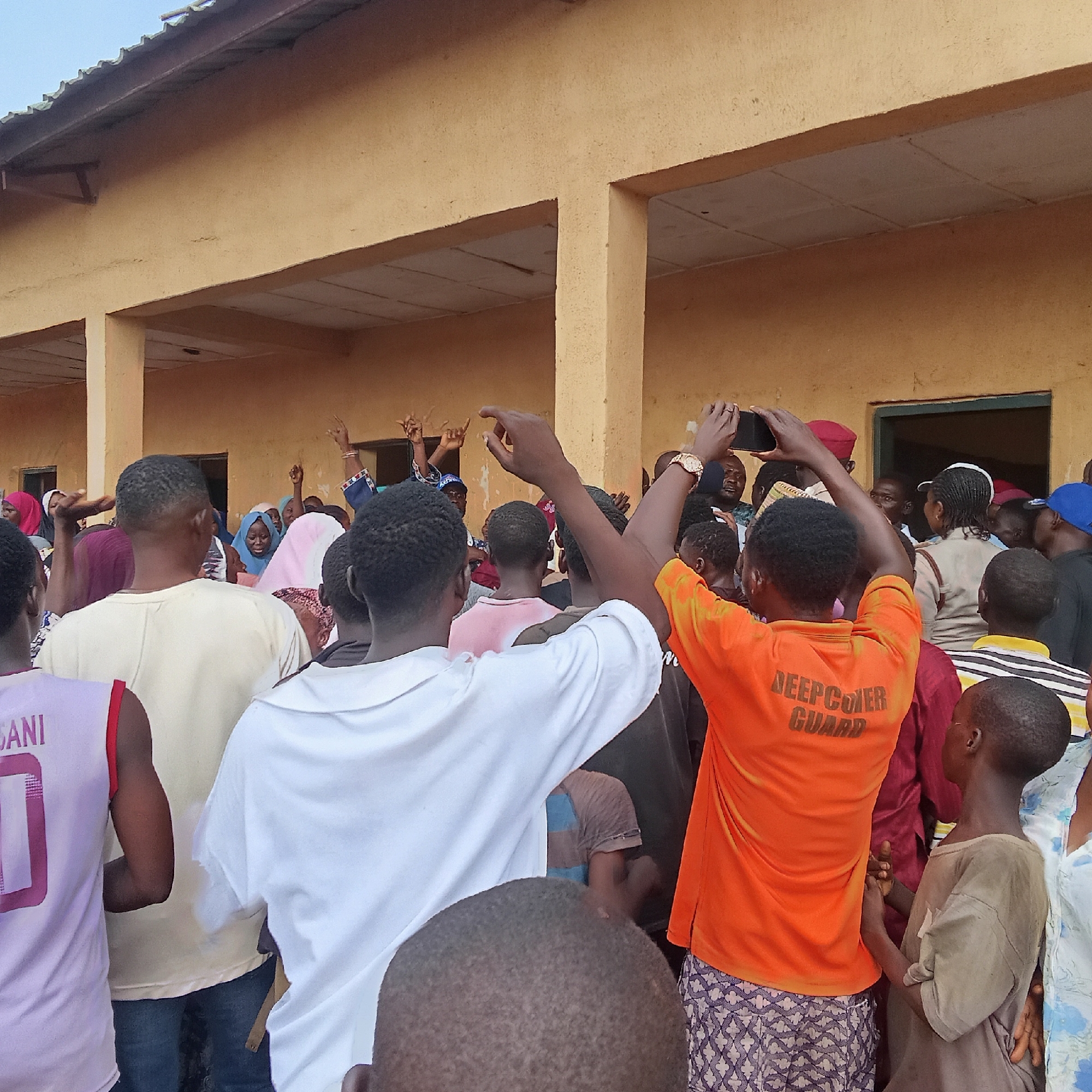 The image shows a crowd of people gathered outdoors in Akwanga, Nigeria. The crowd is in front of a single-story yellow building with a covered veranda supported by columns. Many individuals have their hands raised in the air. A man in an orange shirt with "DEEPCOVER GUARD" printed on the back is holding a smartphone above his head, presumably taking a photo or video. Other individuals are wearing casual attire, including t-shirts, patterned fabrics, and head coverings. The overall atmosphere appears lively.