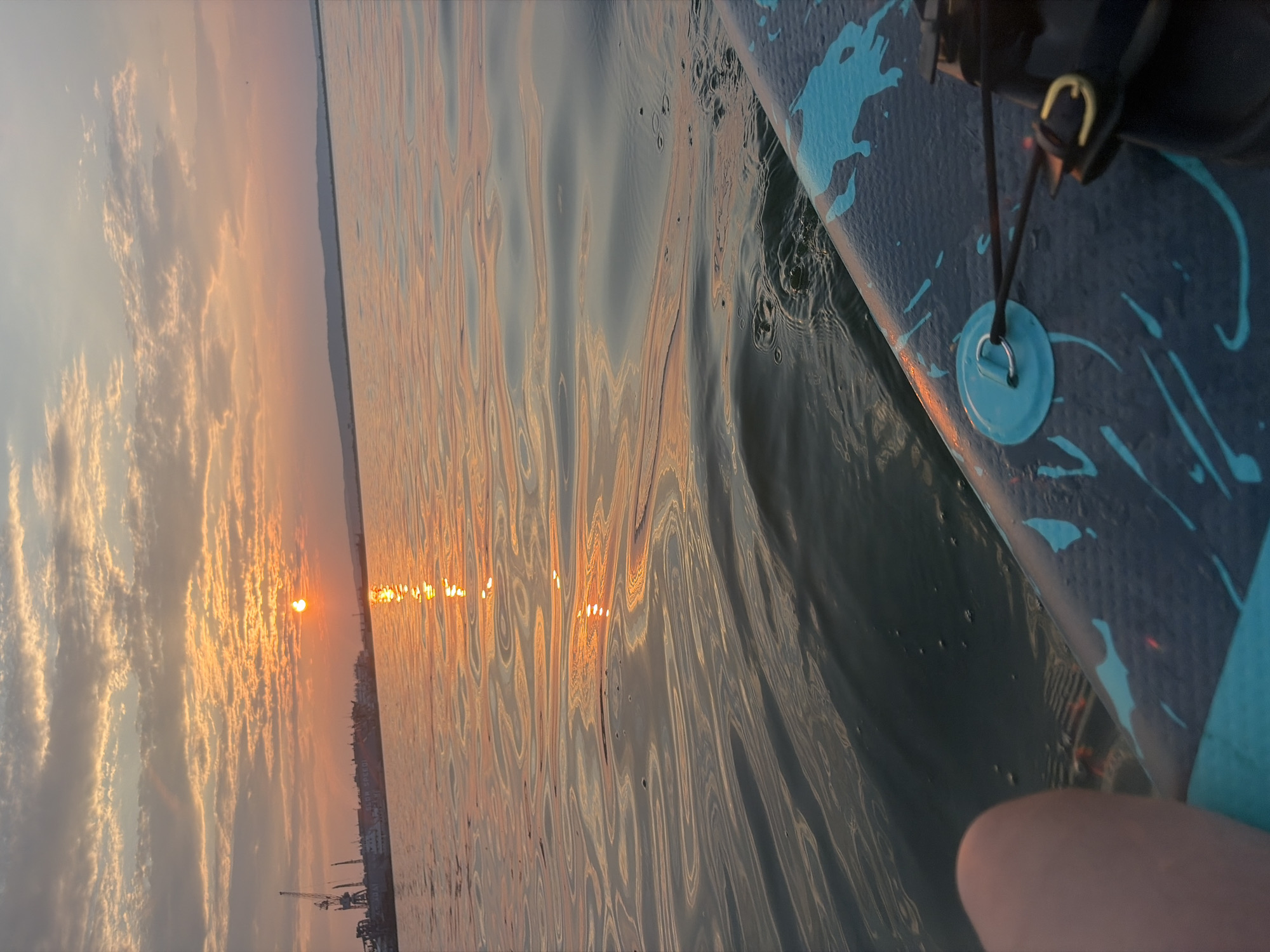 The image depicts a serene scene during sunset, likely on a body of water, viewed from the perspective of someone on a paddleboard or similar watercraft. The sun, a bright orange orb, is setting over a distant shoreline. Wispy clouds are illuminated by the setting sun, casting a warm, golden glow across the sky and reflecting on the rippling water. The water's surface is textured with gentle waves, creating distorted reflections of the sunset.
In the foreground, the edge of a paddleboard is visible, featuring a dark blue surface with turquoise paint splatters. A black rope is attached to a light blue circular fitting with a metal loop. The paddleboard appears to be in motion, indicated by the movement of the water. The distant shoreline shows hints of buildings and industrial structures, possibly cranes, suggesting the presence of a town or port.
The overall mood of the image is peaceful and tranquil, emphasizing the beauty of nature at dusk. The time of day is clearly sunset, given the low angle of the sun and the warm, diffused light. The weather appears calm, with only light ripples on the water. The location is hinted at by the specific mention of Chaikovsky, Russia, which could imply a river or lake setting within that region. There is no visible text in the image.