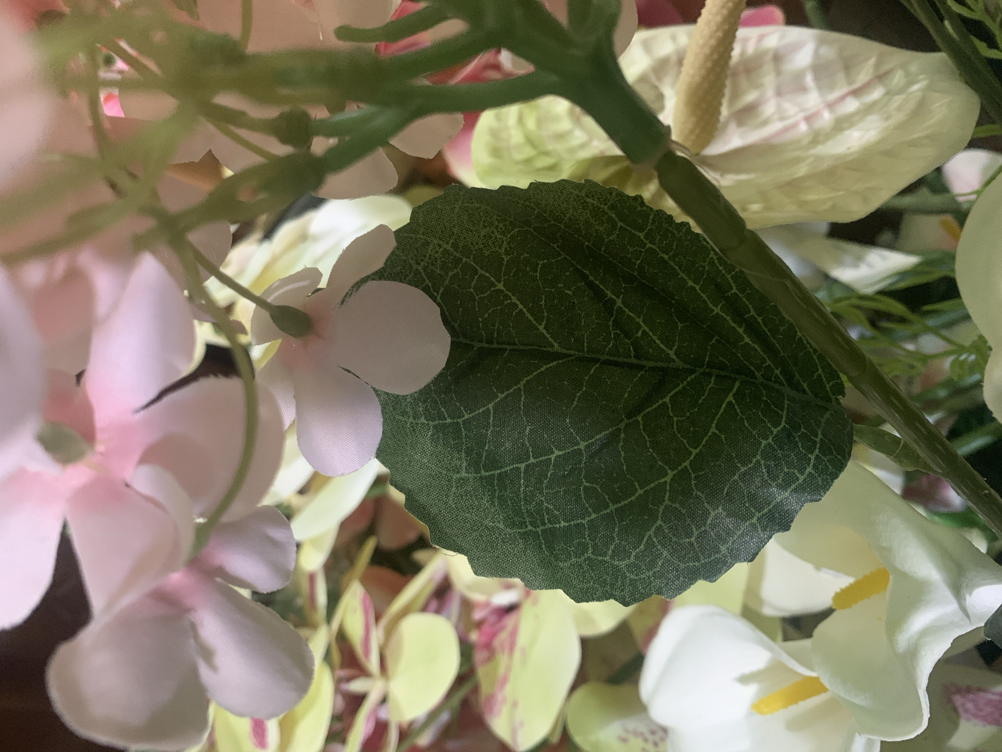 The image is a close-up, macro photograph of artificial flowers. A dark green leaf with a visible veined pattern is prominently featured in the foreground, slightly off-center to the left. Behind the leaf, various artificial flowers in shades of pink, white, and yellow are visible, some in focus and others blurred. The background is dark and indistinct. The scene appears to be a floral arrangement, possibly indoors given the artificial nature of the plants and the lighting. There are no people or animals present. The image does not contain any contextual information that would allow for its location to be identified within Kuala Pembuang, Indonesia, or any other specific geographical location.
