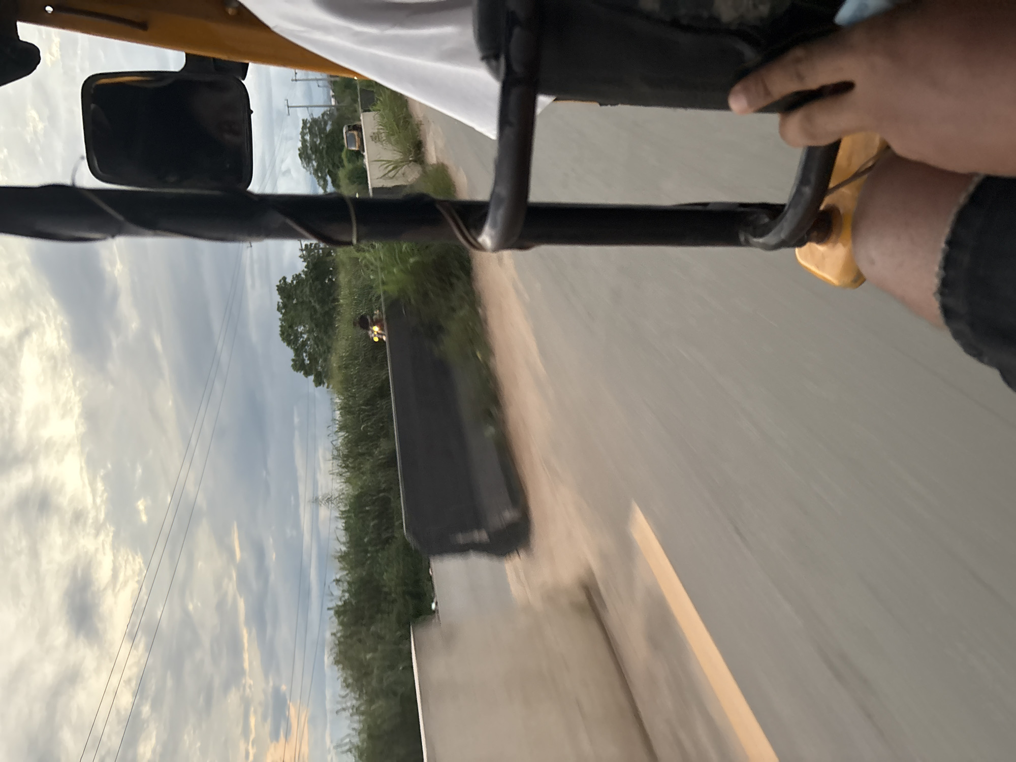 The image shows a view from the perspective of someone riding in a yellow auto-rickshaw in Obosi, Nigeria. The sky is cloudy, with the sun attempting to break through. The road ahead is paved, with a yellow line marking the edge of a lane. To the left of the road, there is a low concrete barrier and dense green vegetation. Further in the distance, more trees and a power line are visible. A motorcycle is seen on the road in the background. The auto-rickshaw has a black metal frame and a yellow body. A person's hand is visible gripping the frame.