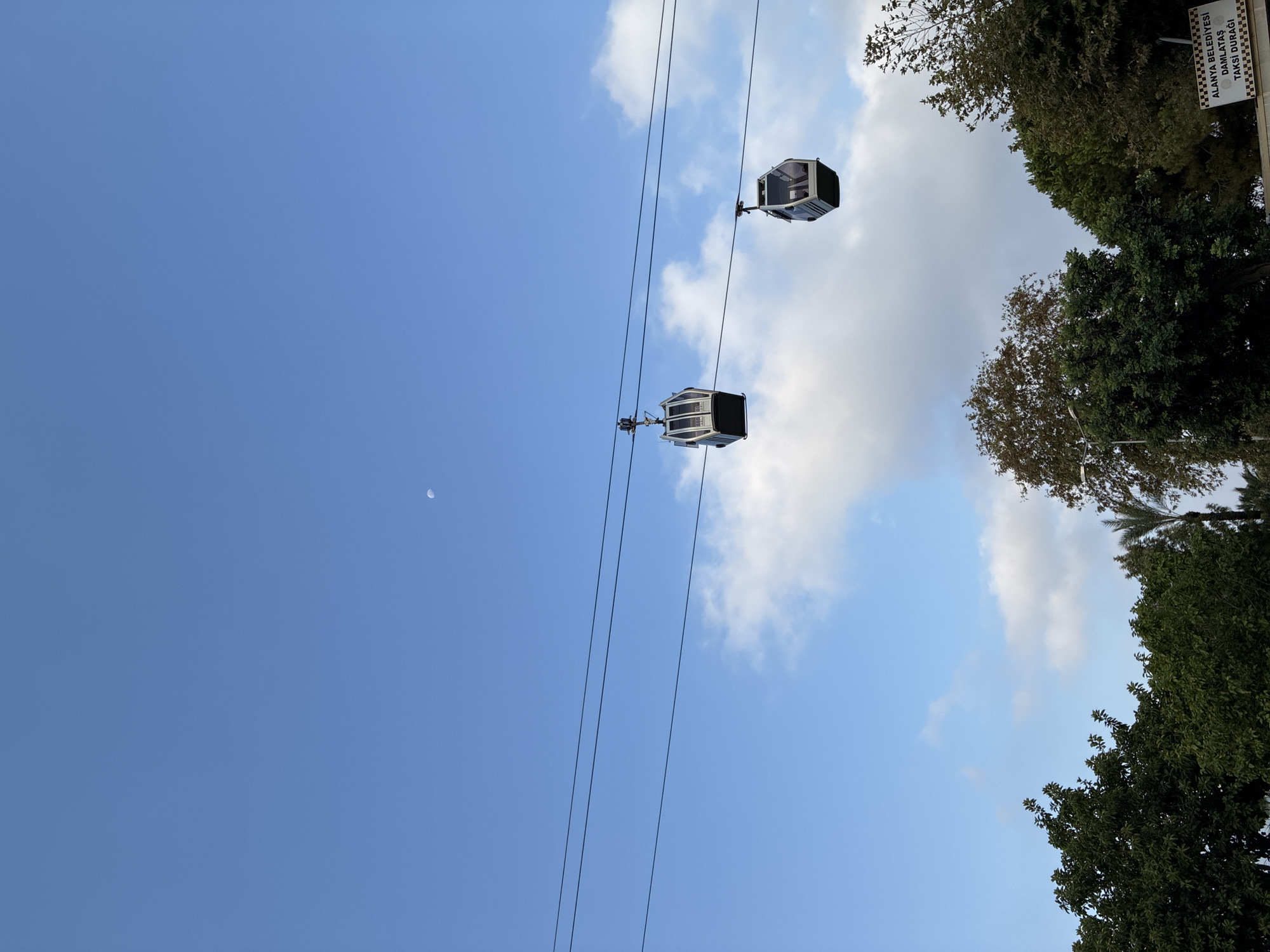 In Alanya, Turkey, two cable car cabins are suspended on parallel wires against a bright blue sky with scattered clouds. The cabins, appearing modern and enclosed with windows, are positioned at different points along the wires. In the upper portion of the sky, a faint crescent moon is visible. The lower part of the image is occupied by lush green trees, with some appearing deciduous and others coniferous. A small sign with text in Turkish is partially visible at the bottom right, identifying a "TAXI STOP" for "ALANYA MUNICIPALITY DAMLATAŞ". The scene depicts an outdoor environment, likely a tourist area, with a focus on the cable car system and natural elements.