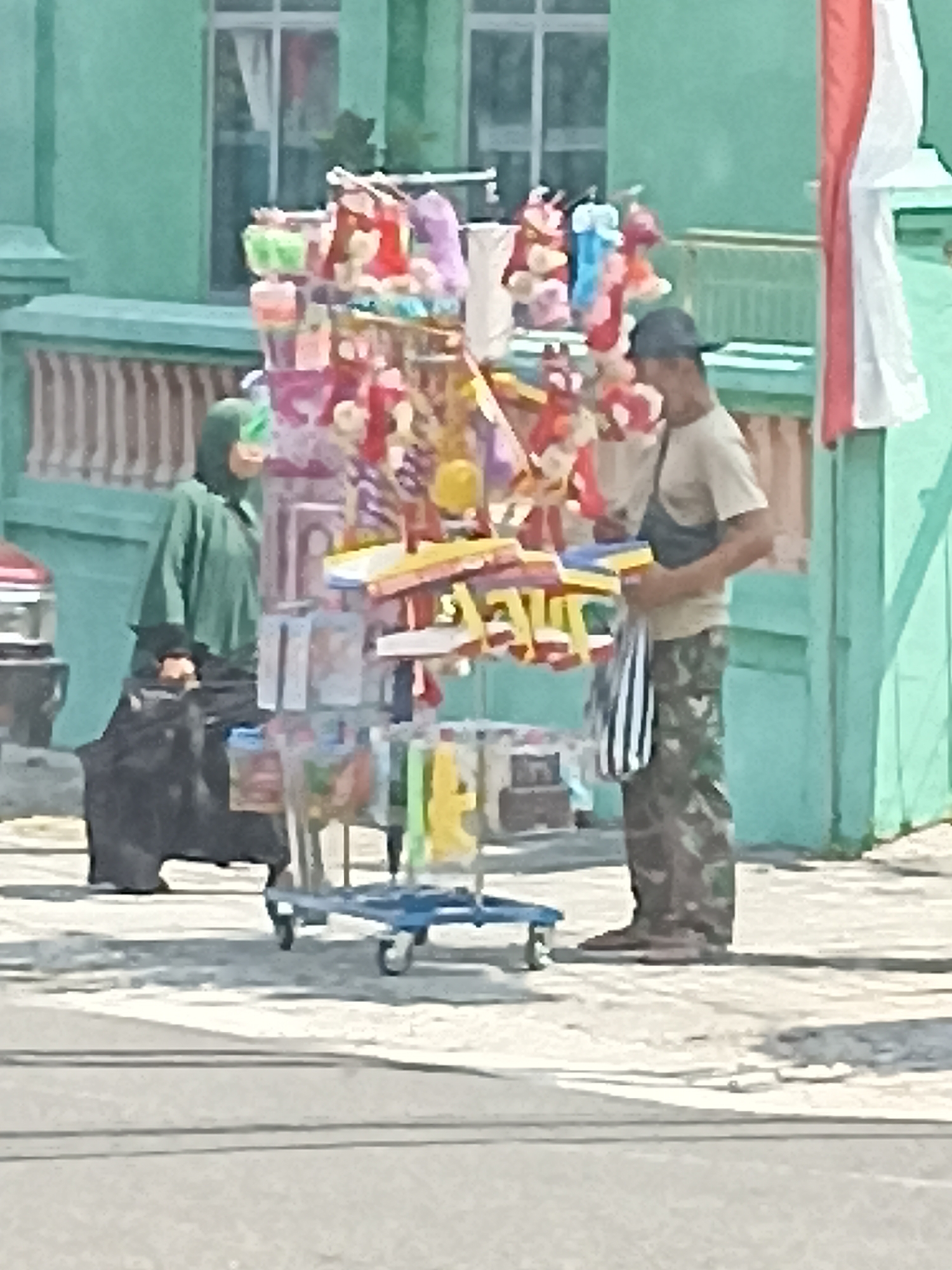 The image captures a street scene in Pulisen, Indonesia, with a focus on a mobile stall selling assorted merchandise. The stall is laden with various items, including what appear to be small stuffed toys and packaged goods, displayed on tiered shelving mounted on a blue wheeled cart. A man wearing a dark cap, a light-colored t-shirt, and camouflage pants is standing next to the stall, holding a striped bag and a yellow object. To the left of the stall, a person wearing a green hijab and dark clothing is standing, with a large black bag at their feet. The background features a light green building with windows and a decorative wall. A red and white flag is visible on the right side of the frame. The ground is paved, and shadows indicate it is daytime. The overall impression is of a lively street vendor scene.