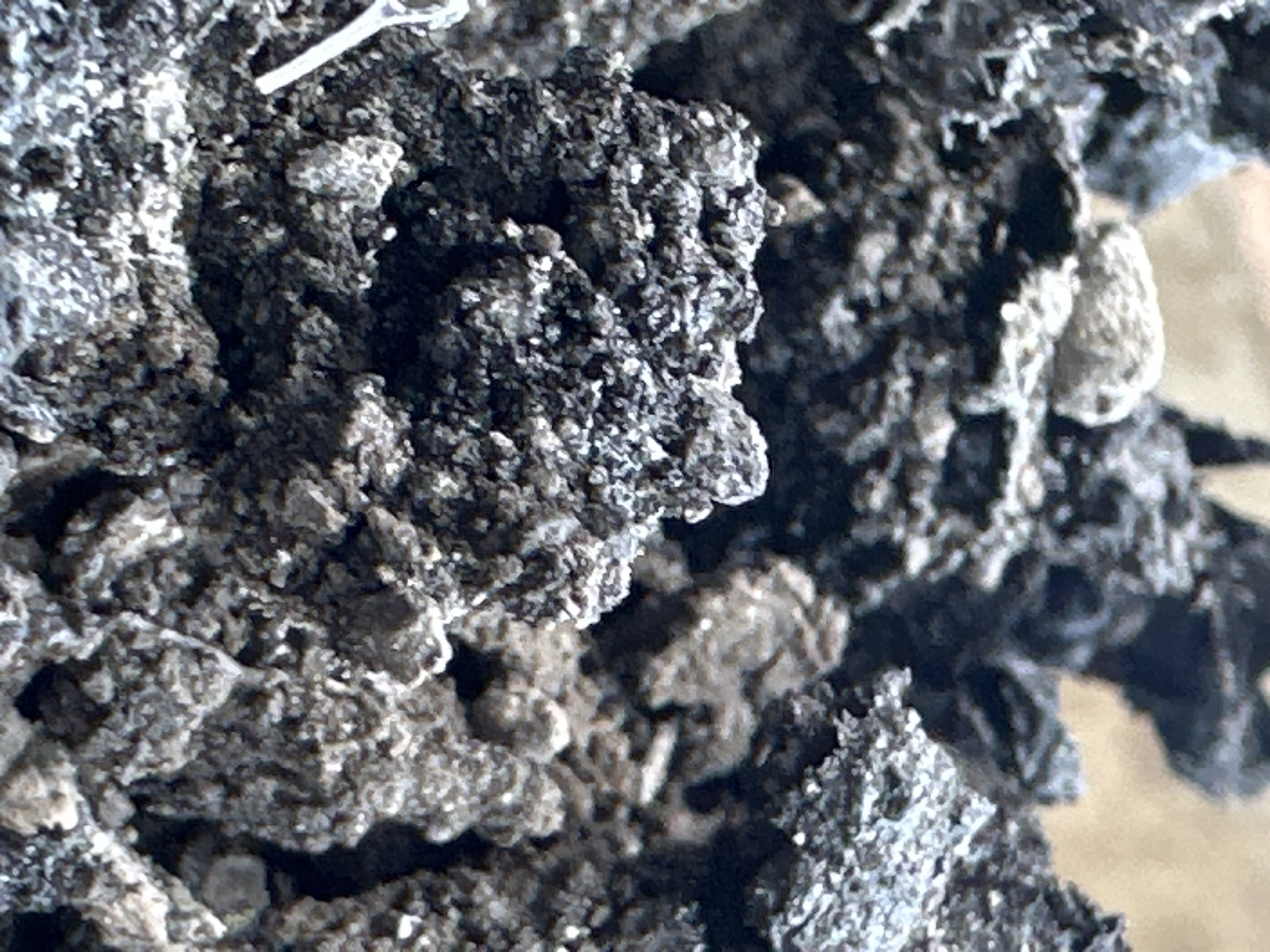 The image is a close-up, macro shot of a pile of dark, granular material, possibly volcanic ash or coarse soil. The texture is rough and uneven, with visible small pebbles and lighter-colored particles interspersed throughout. Some areas appear to be slightly damp, while others are dry and dusty. The background is out of focus, showing a blurred, sandy or earthen tone. No people, actions, or interactions are depicted. The location is specified as Bulakrejo, Indonesia, but the visual content of the image itself does not provide any specific geographical markers or context related to a city.