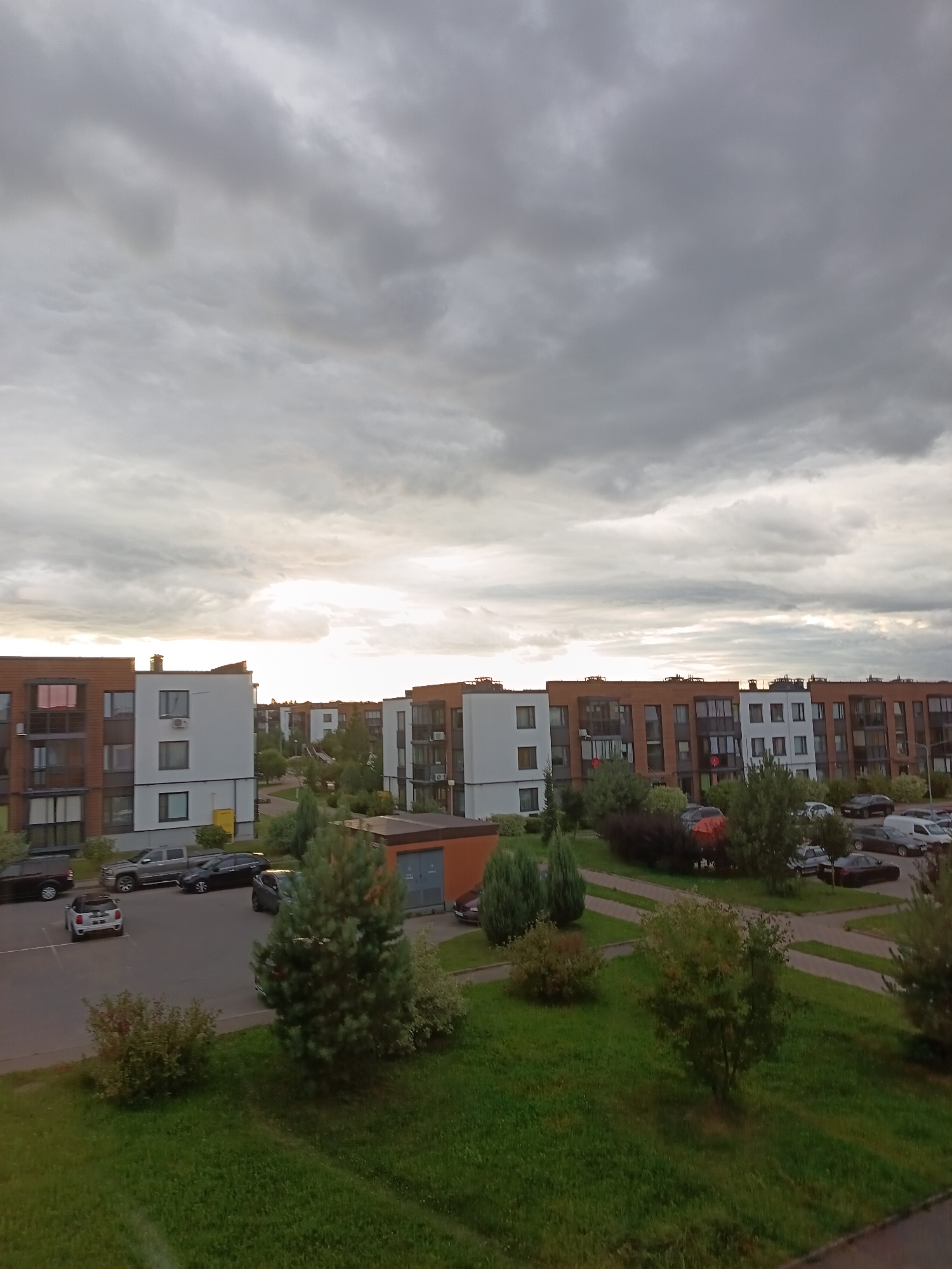 The image shows a residential complex with multiple apartment buildings under a cloudy sky. The buildings are modern, with a mix of white and brown brick facades, balconies, and large windows. There are several cars parked in a lot in front of the buildings, including a silver hatchback, a dark sedan, and a grey pickup truck. A small evergreen tree is prominently featured in the foreground. The scene appears to be a typical suburban or semi-urban environment, likely captured during the daytime given the overall illumination. There are no people visible in the image. The sky is overcast with dramatic cloud formations.
