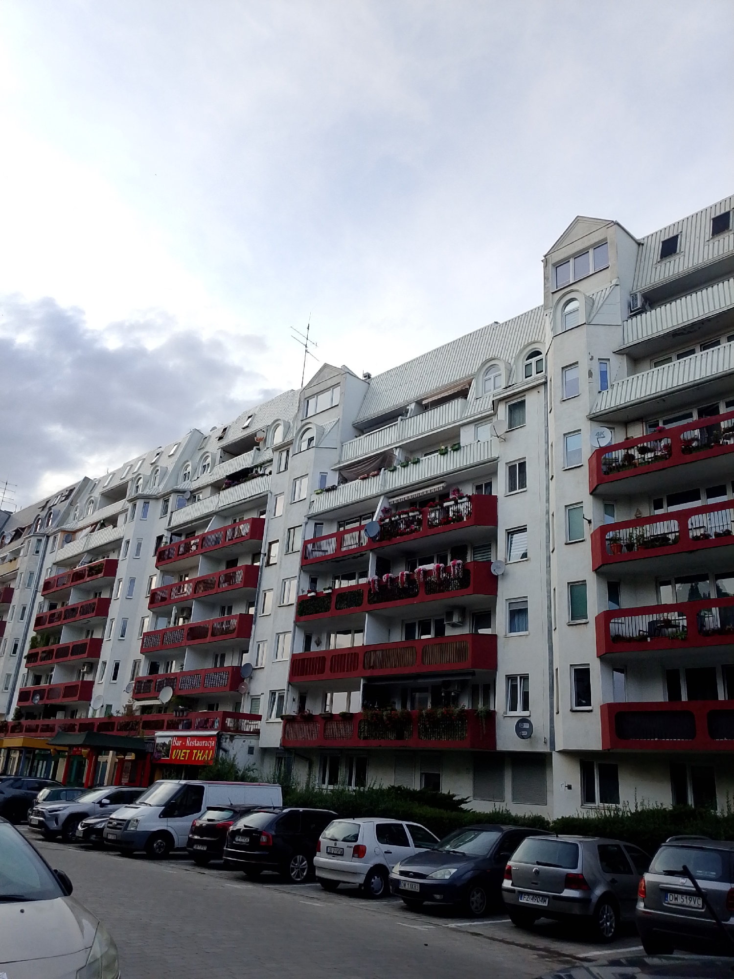 The image shows a residential building with multiple floors and many balconies. The building is painted white with red railings on the balconies. There are several cars parked in front of the building, and some greenery is visible. The sky is cloudy, and the overall lighting suggests it is daytime. A sign on the building reads "Bar - Restauracja VIET THAI." The building is located in Wrocław, Poland.
