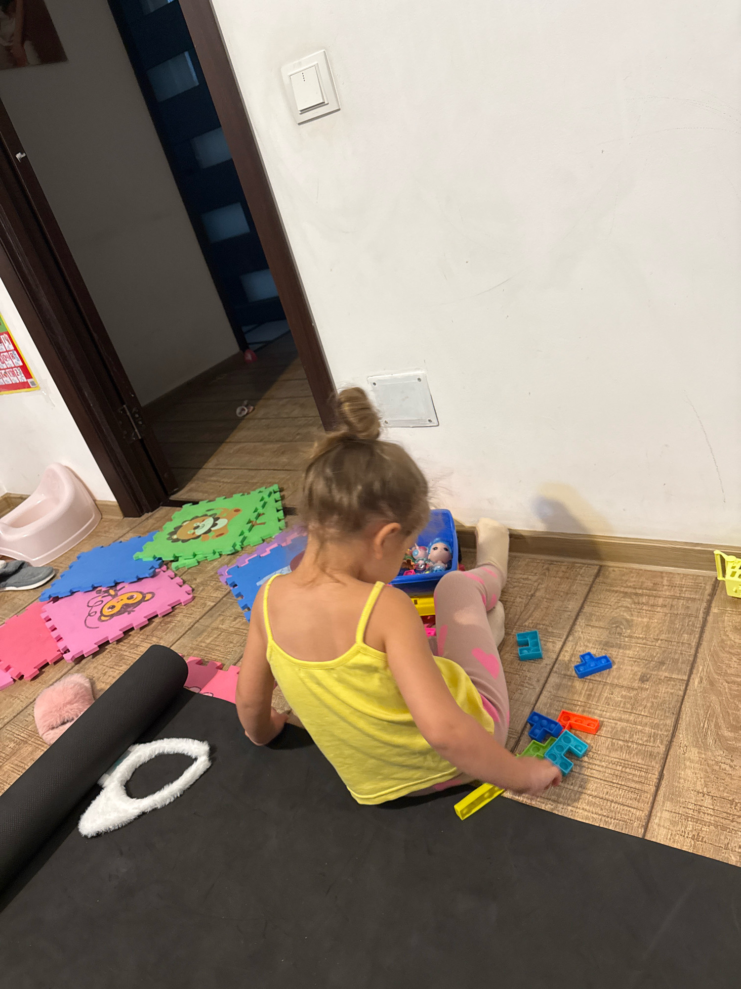 A young girl with blonde hair in a bun is sitting on a wooden floor in a room, playing with colorful interlocking toys. She is wearing a yellow tank top and pink leggings with heart patterns. Her back is to the camera as she reaches for the toys. Scattered around her are several foam play mats with cartoon animal designs, a rolled-up black yoga mat, and a pink fluffy slipper. In the background, a doorway reveals a dark hallway, and a white light switch is visible on the wall. There is also a light pink toddler potty near the doorway. The overall scene depicts a child engaged in play within a home environment, likely in Chișinău, Moldova, based on the provided location context.