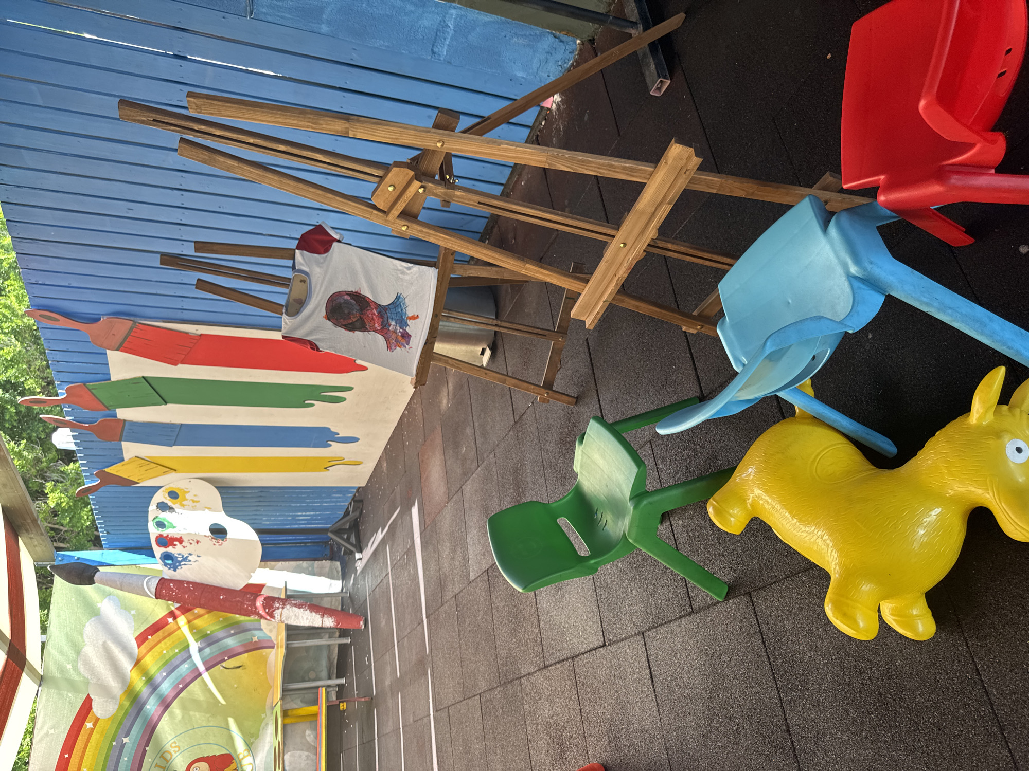 The image depicts an outdoor art area, likely a children's art class or play area, possibly located in Aksu, Turkey, given the context of the inquiry. The scene is set against a bright blue wooden fence adorned with oversized, painted representations of paintbrushes in red, yellow, blue, and green. In the background, a large mural features a rainbow and clouds.
In the foreground, a series of child-sized plastic chairs are scattered on a dark, rubberized floor. A bright green chair is prominently positioned. Nearby, a bright yellow inflatable animal, possibly a horse or a similar toy, lies on its side. A light blue chair and a red chair are also visible.
To the right of the scene, two wooden easels stand. One easel displays a white t-shirt with a printed image of a person's head wearing a Spider-Man mask. The other easel is empty. A large, oversized palette with splotches of colorful paint is mounted on the blue fence, next to a large, red-handled paintbrush with white paint residue. A yellow table with red legs is partially visible in the left background. The overall impression is a lively, colorful space designed for creative activities.