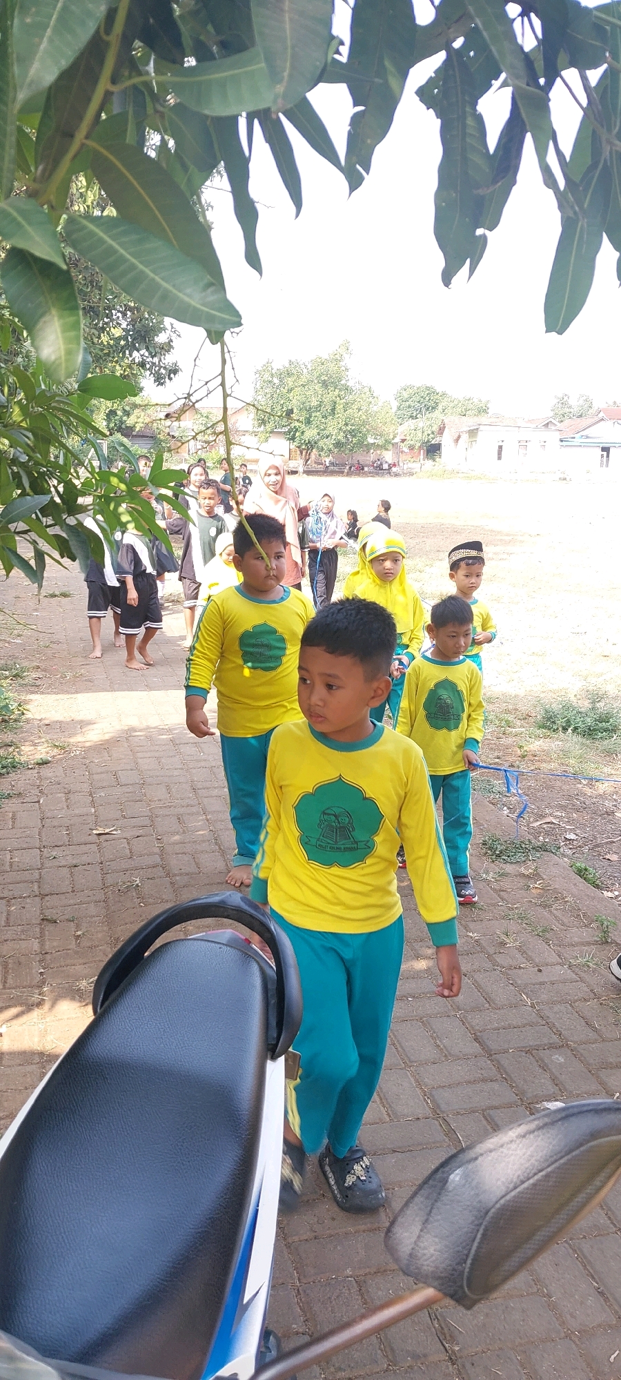 In Keling, Indonesia, a group of children in yellow shirts and teal pants walks along a paved brick path under the shade of mango tree leaves. The children appear to be participating in an outdoor activity or procession, with some wearing headscarves. In the foreground, the seat and handlebars of a motorcycle are visible, partially obscuring the path. The background shows residential buildings and trees, suggesting a suburban or village setting. The scene is bathed in bright sunlight, indicating a clear day.