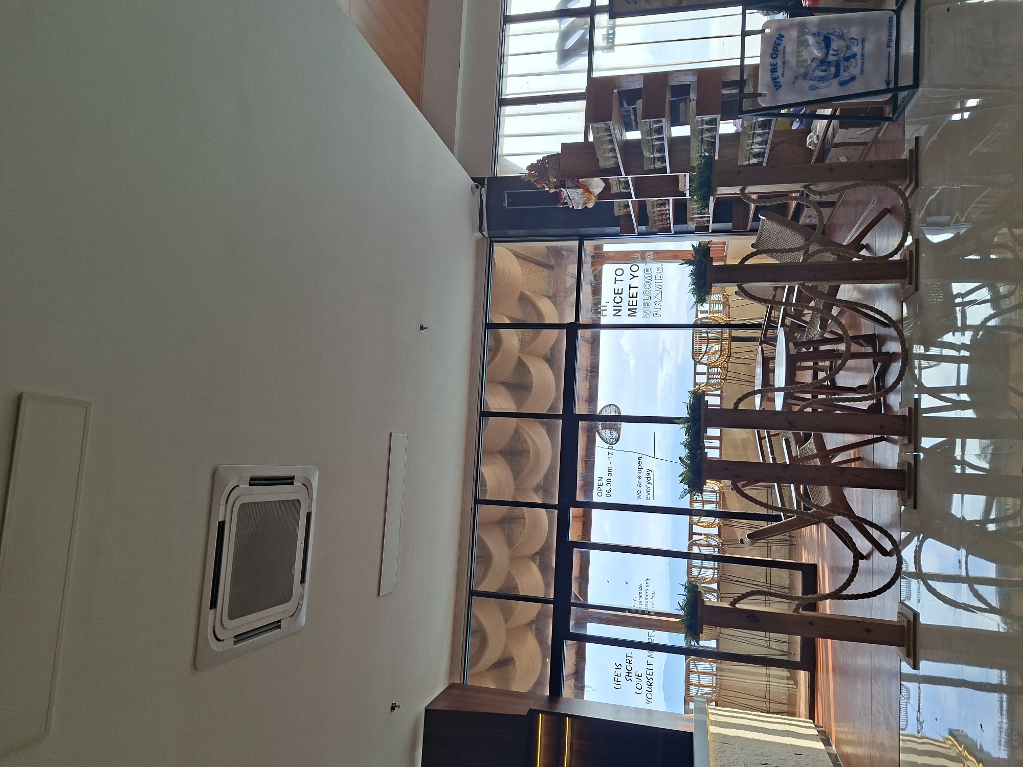 This appears to be an interior view of a cafe or restaurant in Nusa Penida, Indonesia. The space is well-lit, and the focus is on the dining area near a large window. Tables and chairs are arranged, with the chairs having a woven or rope design. Plants are positioned atop the table stands. Sunlight streams through the glass. Window advertisements read “WE'RE OPEN COFFEE AND PASTRY”, "OPEN 06.00 AM-17.00 PM", and “HI, NICE TO MEET YOU. WELCOME TO PIRAMIDE. LIFE IS SHORT. LOVE YOURSELF MORE”. There are shelves visible near the window. The interior is modern and well-maintained. An air conditioning vent is visible on the ceiling.