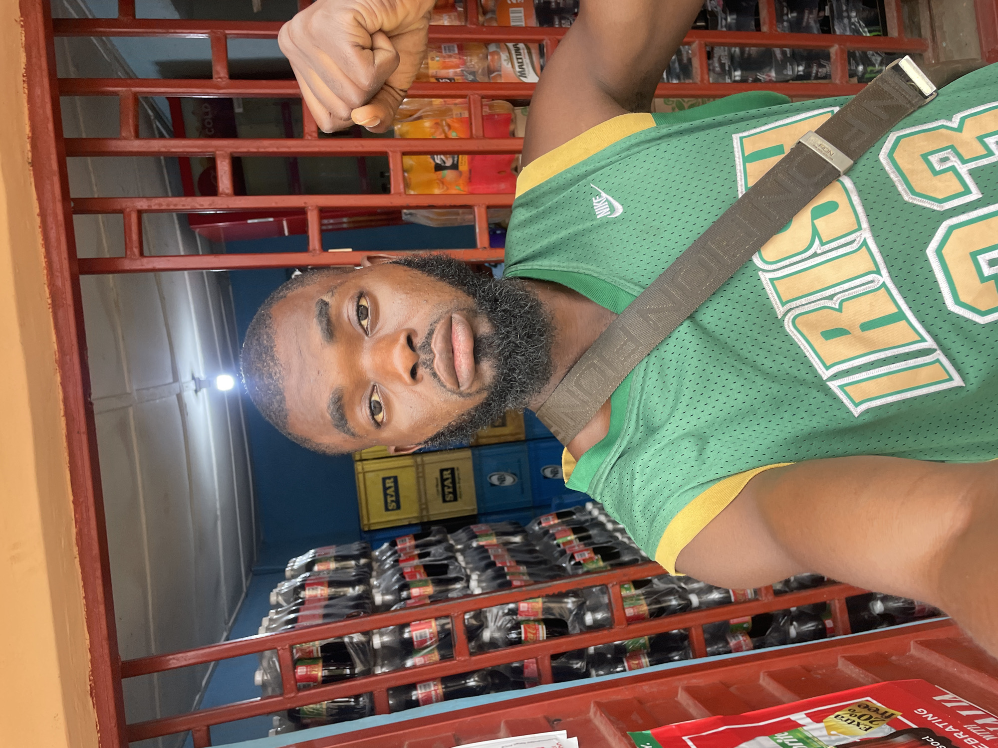 The image depicts a man posing for a selfie in front of a store with shelves stocked with beverages, likely in Jos, Nigeria. The man is wearing a green basketball jersey with yellow trim and the word "IRISH" and the number "23" visible. He has a beard and is wearing a crossbody bag. Behind him, bottles of Coca-Cola and other drinks are stacked high on metal shelves. Yellow and blue crates are also visible in the background, some with "STAR" branding. The store appears to be an open-air or semi-open space, with a red metal gate structure in the foreground and a white ceiling with lighting. The overall impression is of a retail environment selling drinks.